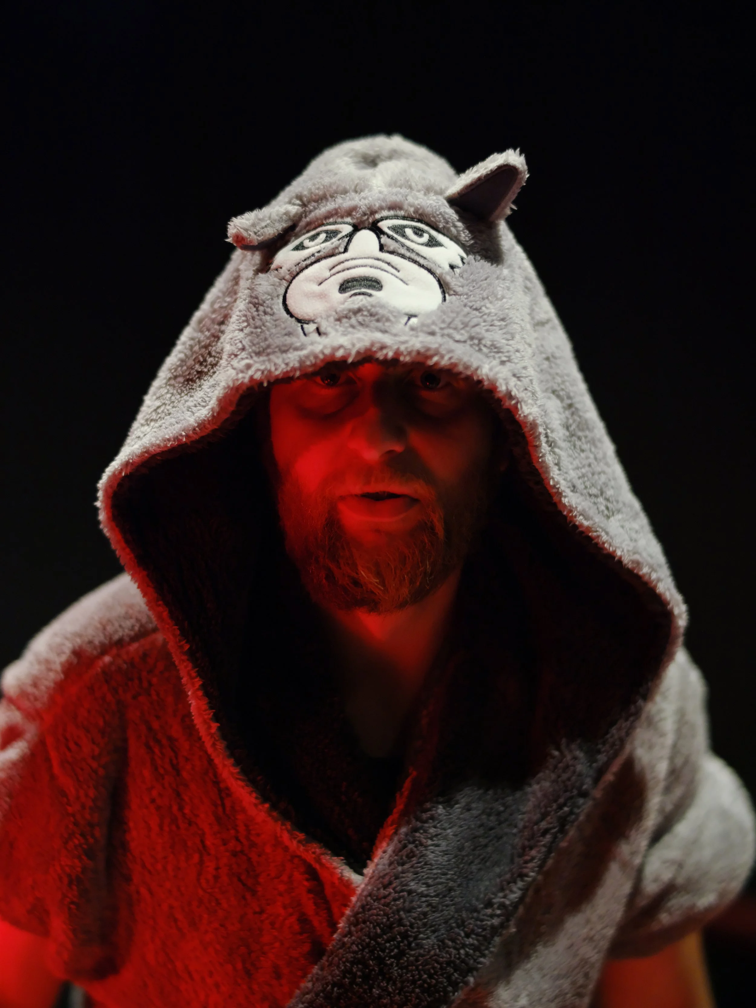 A person wearing a hoodie with a cartoon face embroidered on it, with a distressed expression, and a character with glasses and a beard on the hood. The person is illuminated with red light in a dark setting.