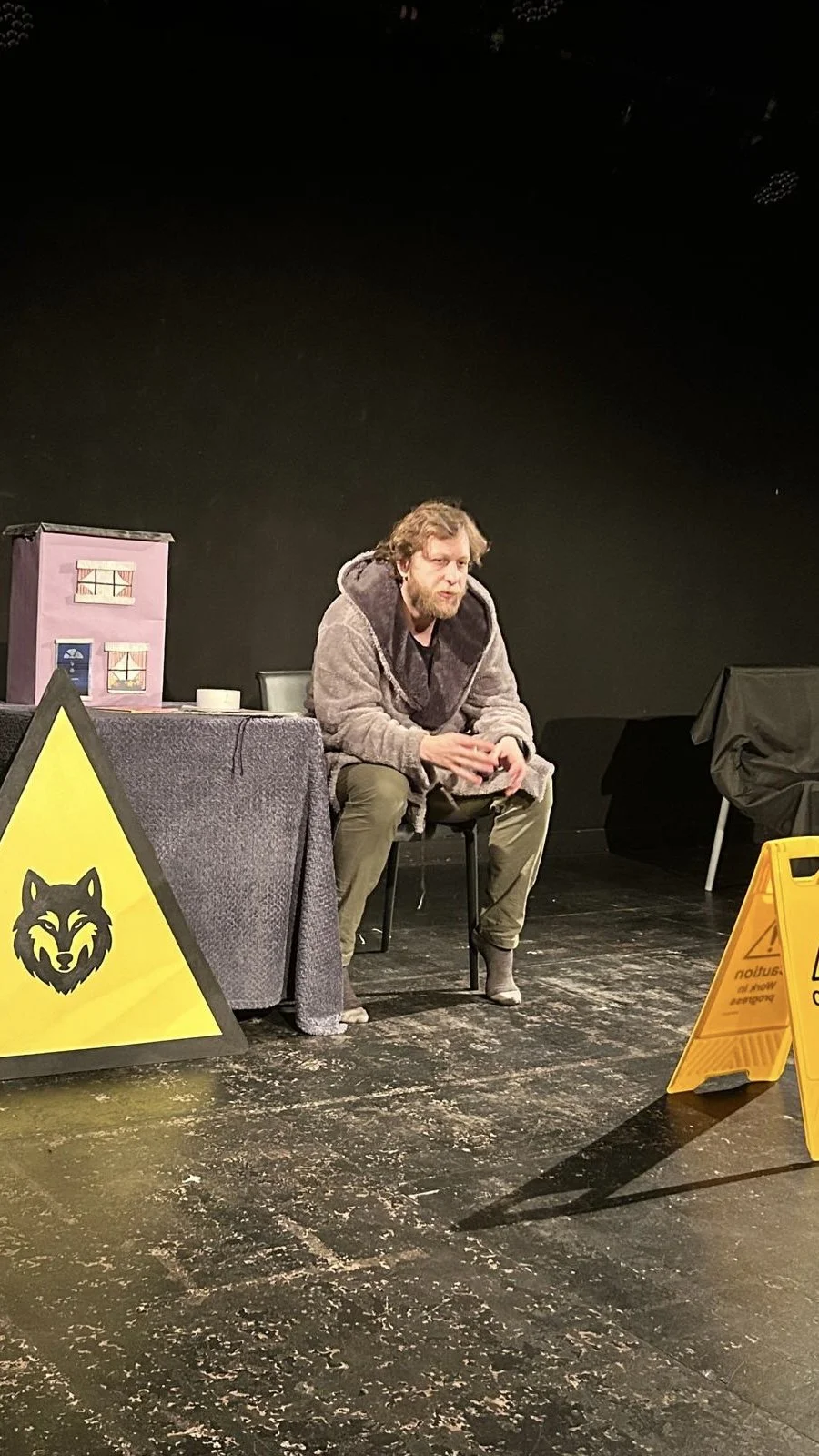 A man sitting on a small stool on a stage, wearing casual clothes and a hoodie, with a serious expression. There are signs on the floor indicating wet floor or caution, and a black backdrop, with some props and furniture around.