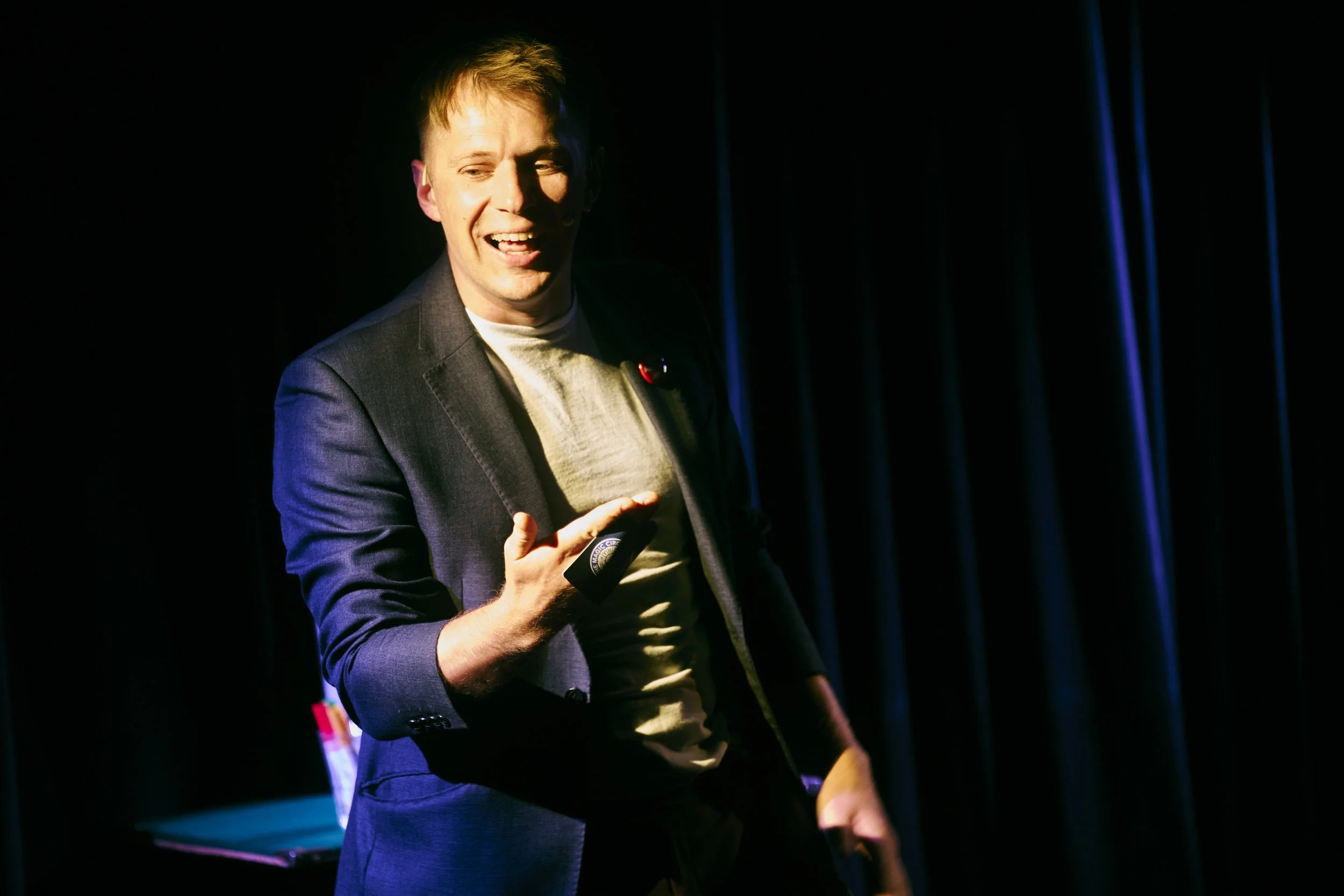 A young man in a blazer on stage, speaking into a microphone, with a dark background and blue curtains.