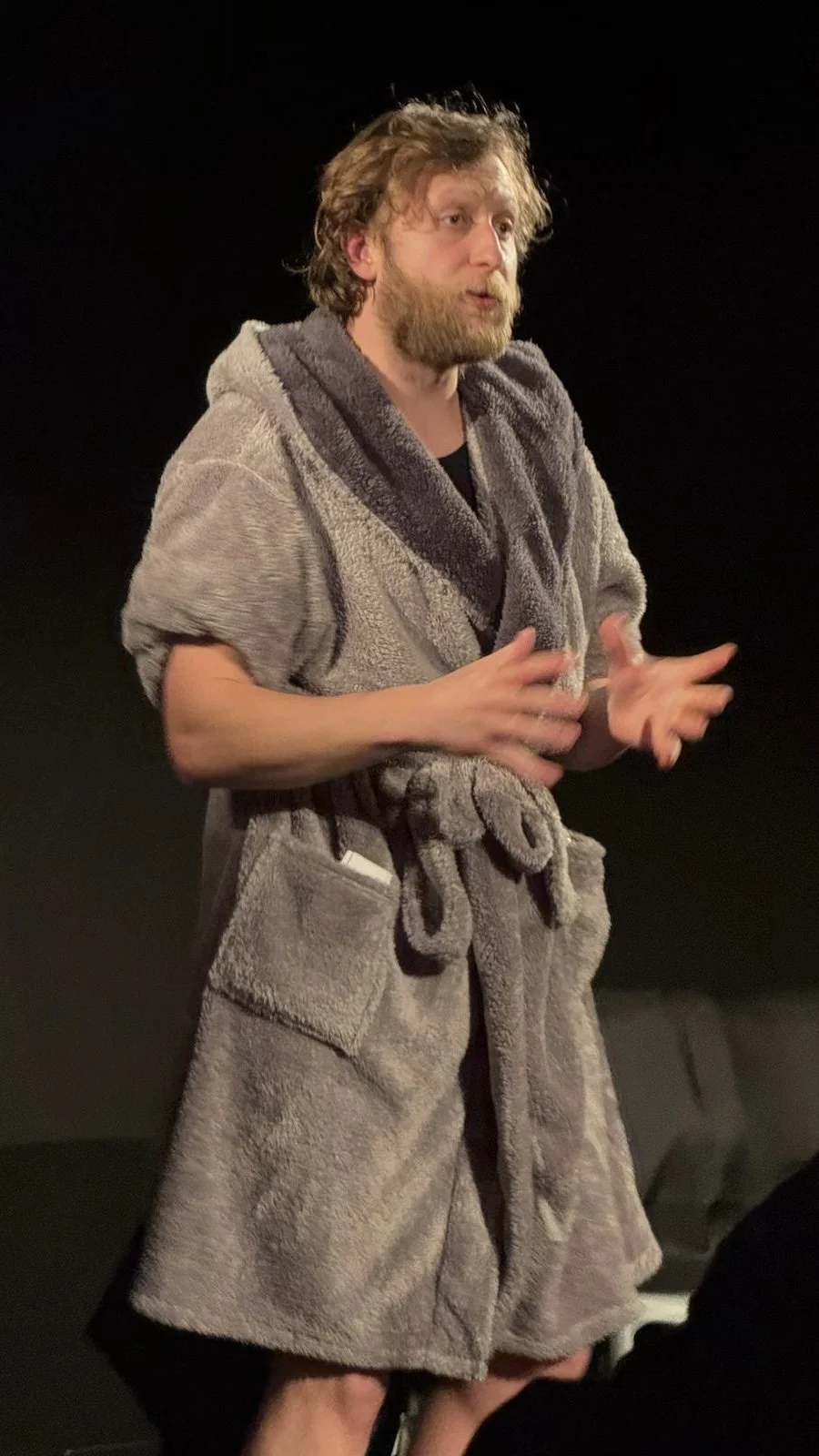 A man with a beard and disheveled hair is wearing a large, fluffy bathrobe and gesturing with his hands, standing against a dark background.