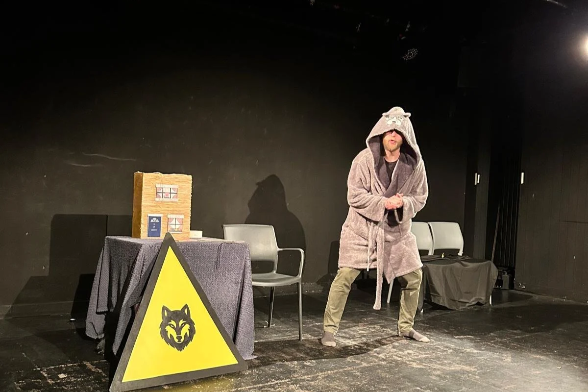 A man dressed in a bear costume with a hood featuring a bear face, standing on a small stage in a theater. There is a table with a black tablecloth, a cardboard box, and chairs on stage, with a yellow warning sign with a wolf head symbol leaning agai