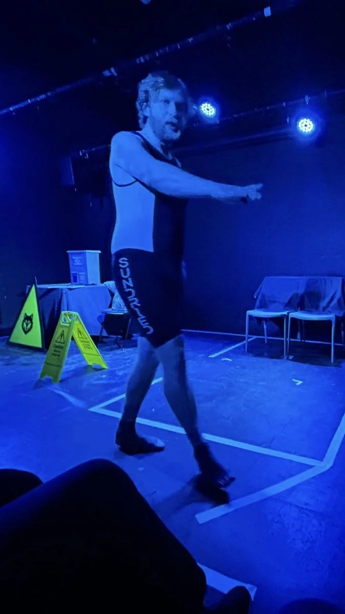 A man standing in a dark room with blue lighting, wearing black shorts with the word 'SUNDAYS' on the side, possibly performing or practicing a dance or exercise.