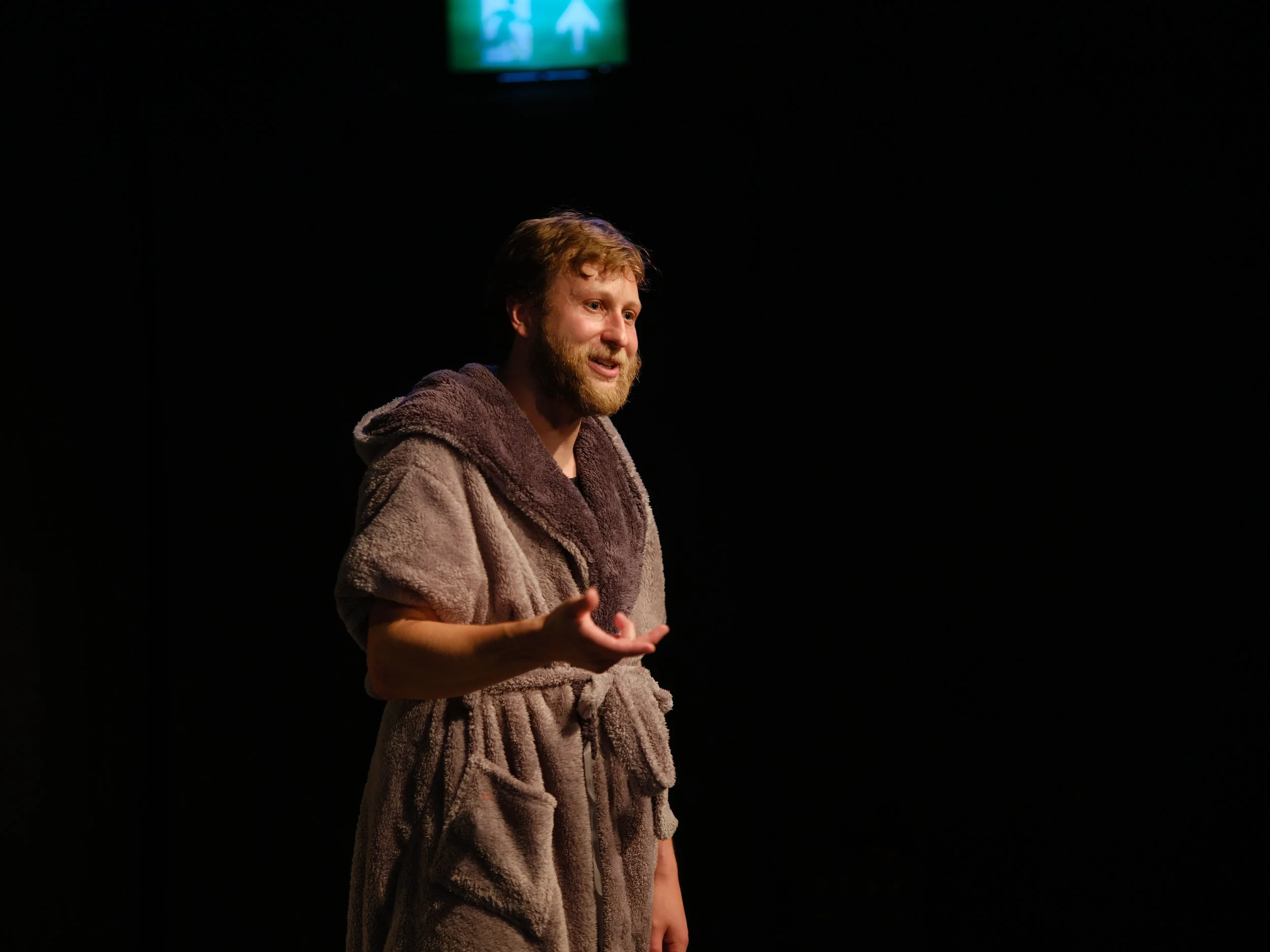 A man with a beard and curly hair wearing a brown fuzzy coat is speaking or performing on a dark stage with a green exit sign above him.