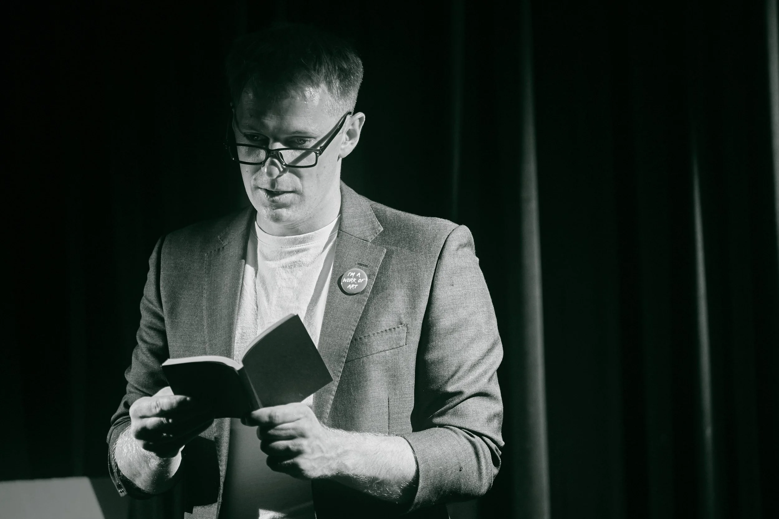 Man wearing glasses, reading a book on stage, wearing a blazer with a button that says 'I'M A WORK OF ART', with dark curtains in the background.