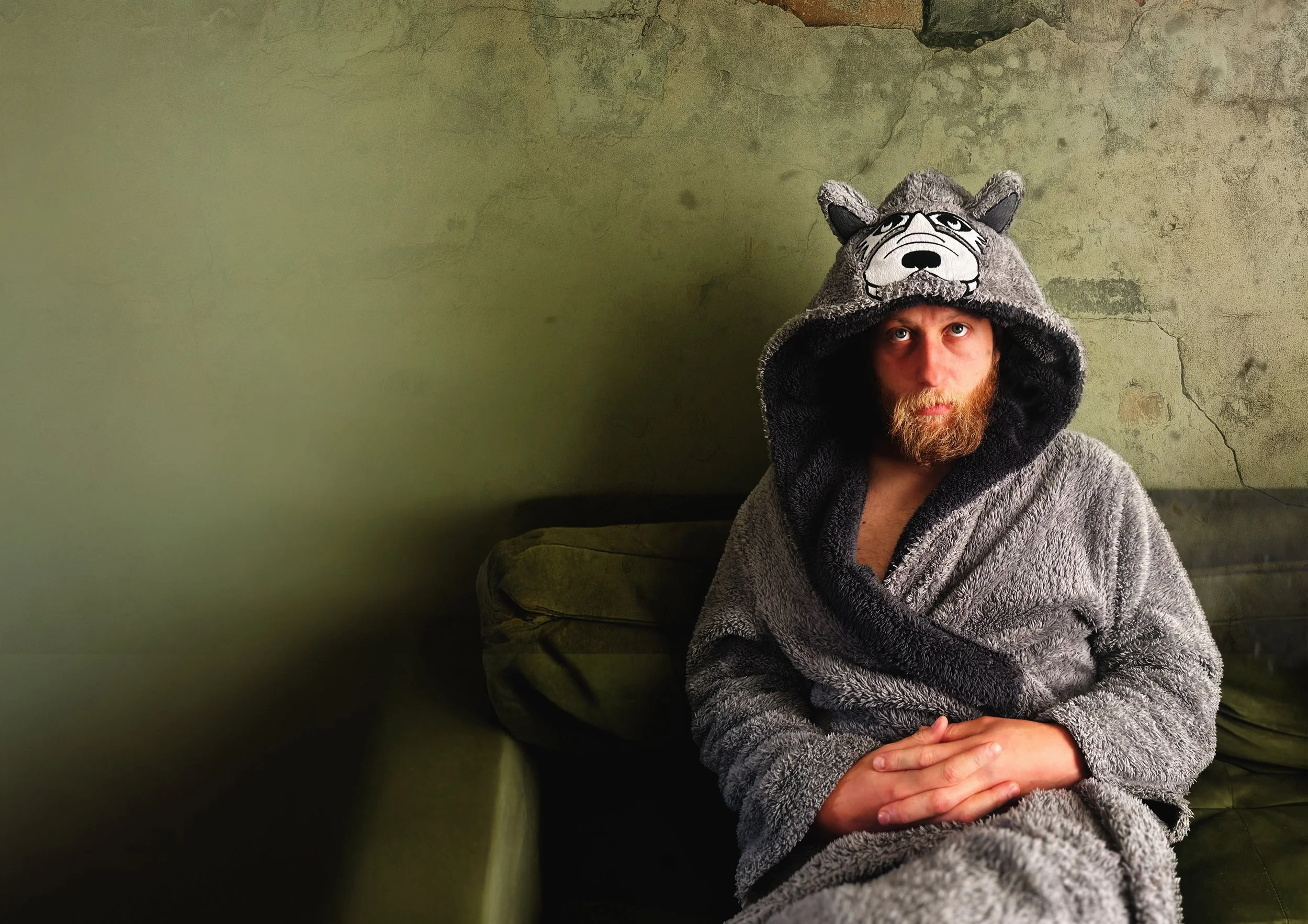 A person with a beard wearing a hooded bathrobe is sitting against a textured, greenish wall. They are looking upward, with hands folded on their lap, and are wearing a gray furry animal-themed hat resembling a wolf.