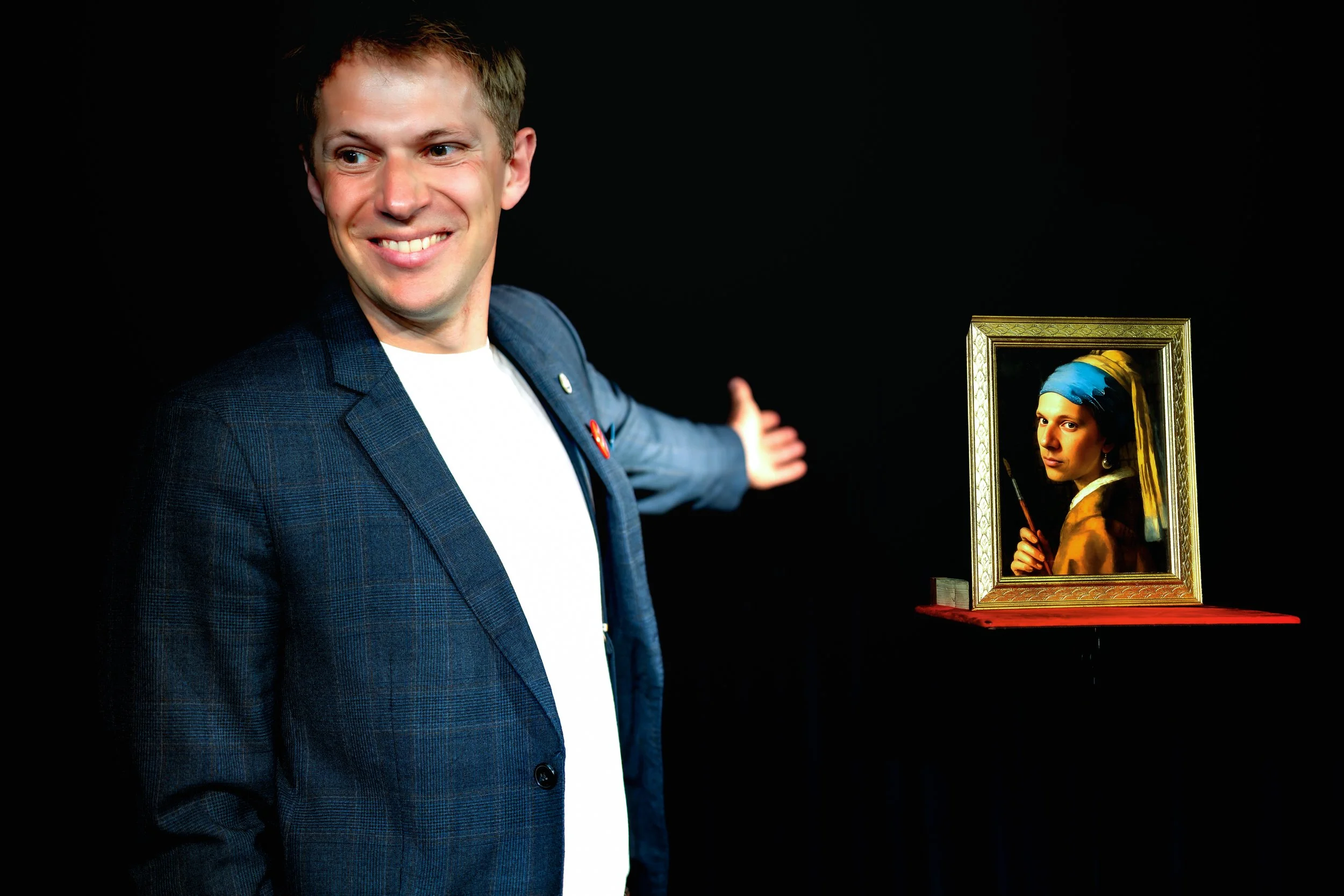 A man smiling and gesturing towards a framed painting of a woman in traditional attire, displayed on a table against a black background.