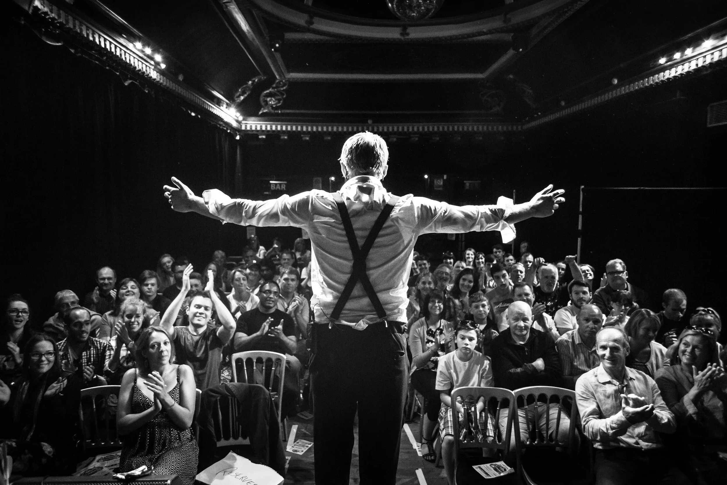 A performer dressed in a white shirt with suspenders stands on stage with arms outstretched, facing a large audience in a theater. The audience is clapping and smiling, and the theater has ornate ceiling details with dim lighting.