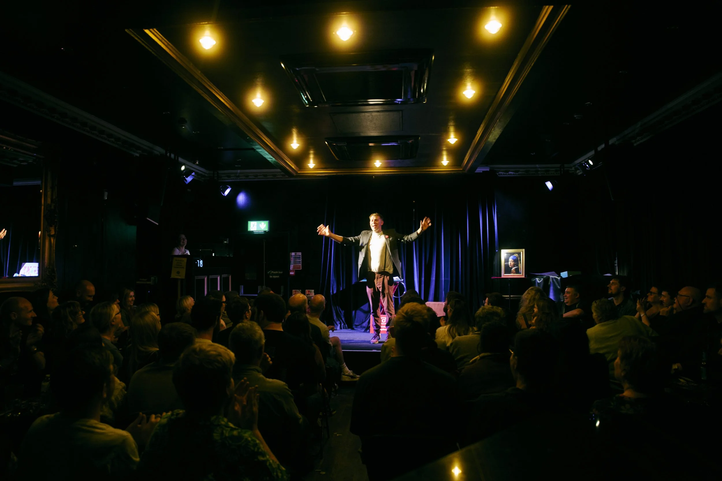 A male performer stands on a stage with his arms outstretched, engaging with an audience in a dark theater. The crowd watches attentively, and stage lighting illuminates the performer and the surroundings.
