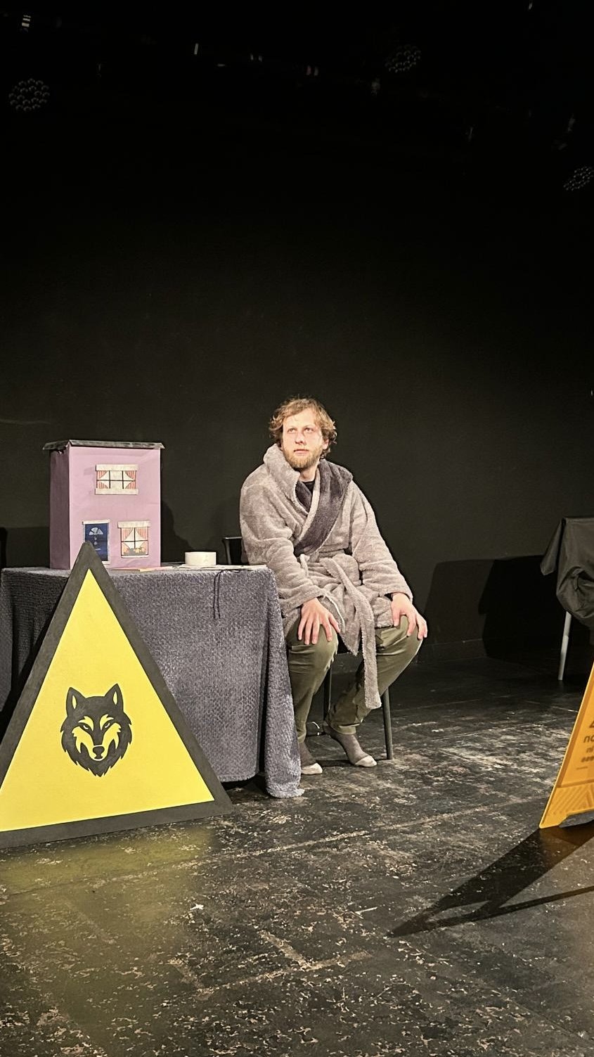 A man in a bathrobe sitting on a chair on a stage with a black backdrop. There are signs and props, including a yellow warning sign with a wolf's face, a purple box with windows, and a yellow caution sign with writing.