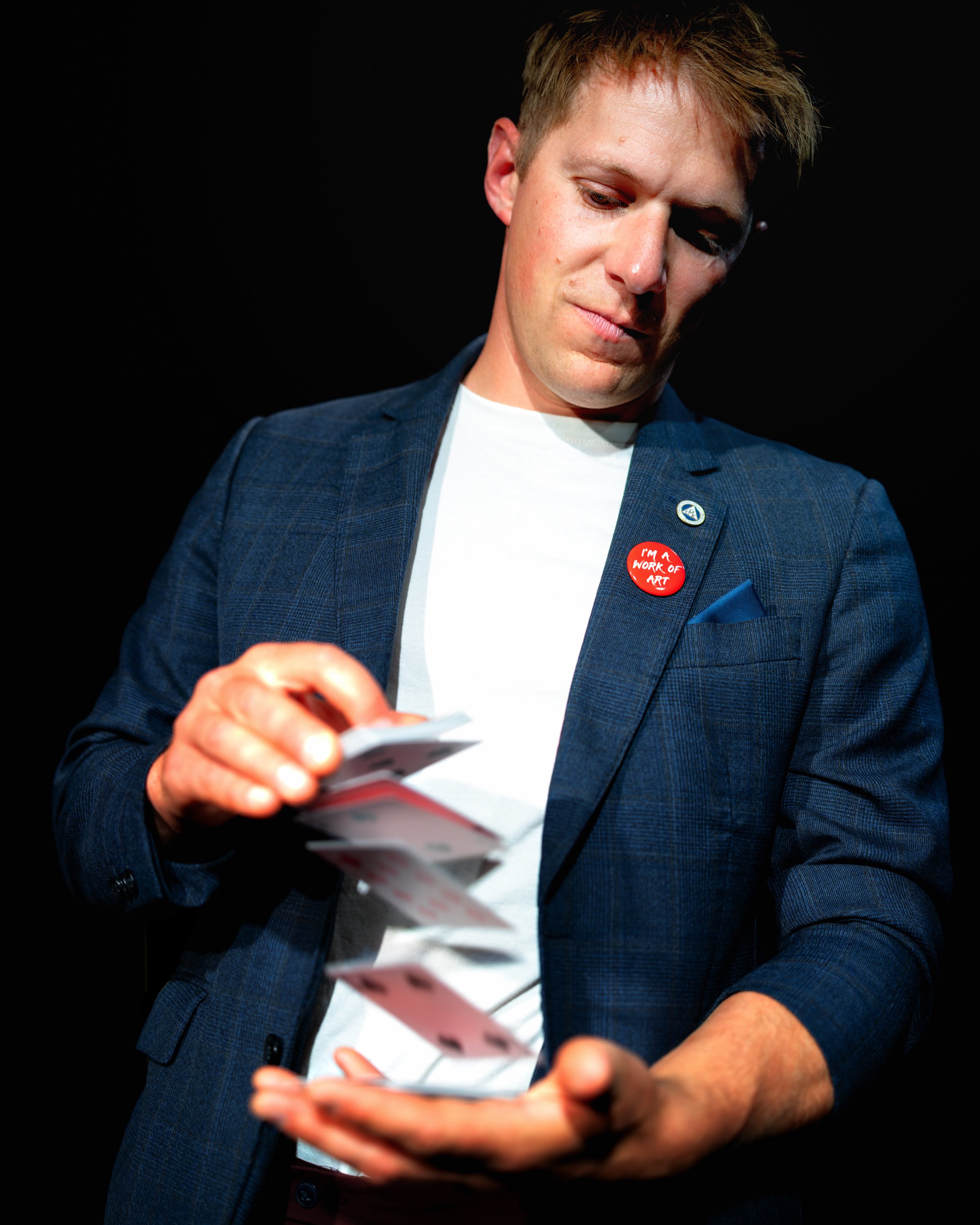 A young man wearing a dark blue blazer and a white shirt, looking down at a deck of cards he's holding, with a red button that reads 'I'm a work of art' pinned to his blazer. The background is black.