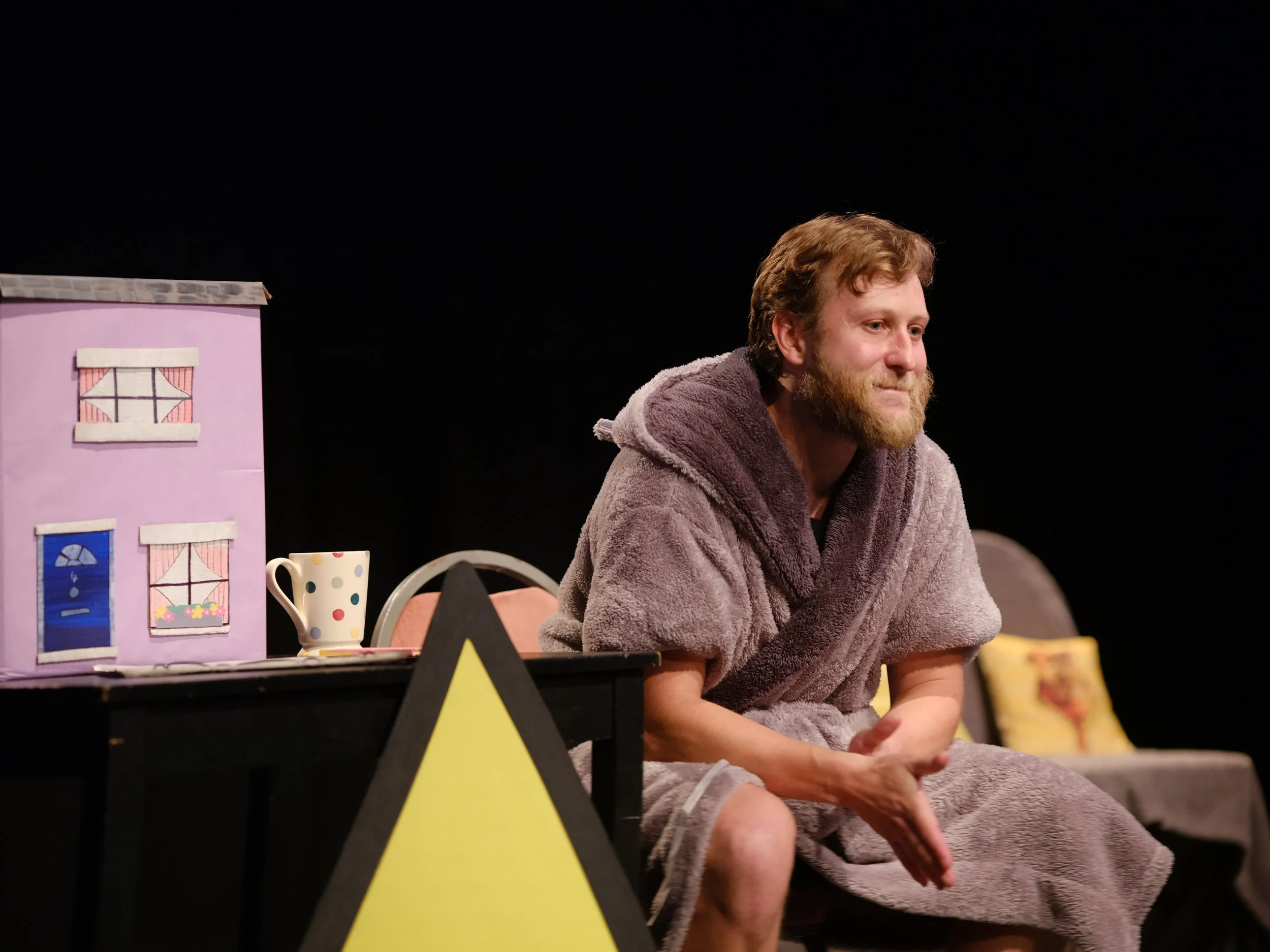 A man with a beard, wearing a brown bathrobe, is sitting on stage with theatrical props including a pink dollhouse and a polka dot mug. The background is black.
