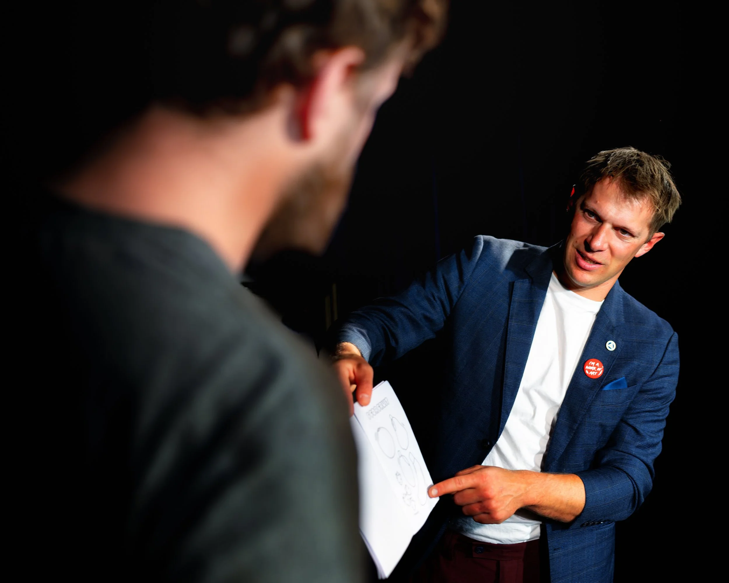 Two men engaging in conversation, one wearing a blue blazer and the other with a blurred face, in a dark setting.