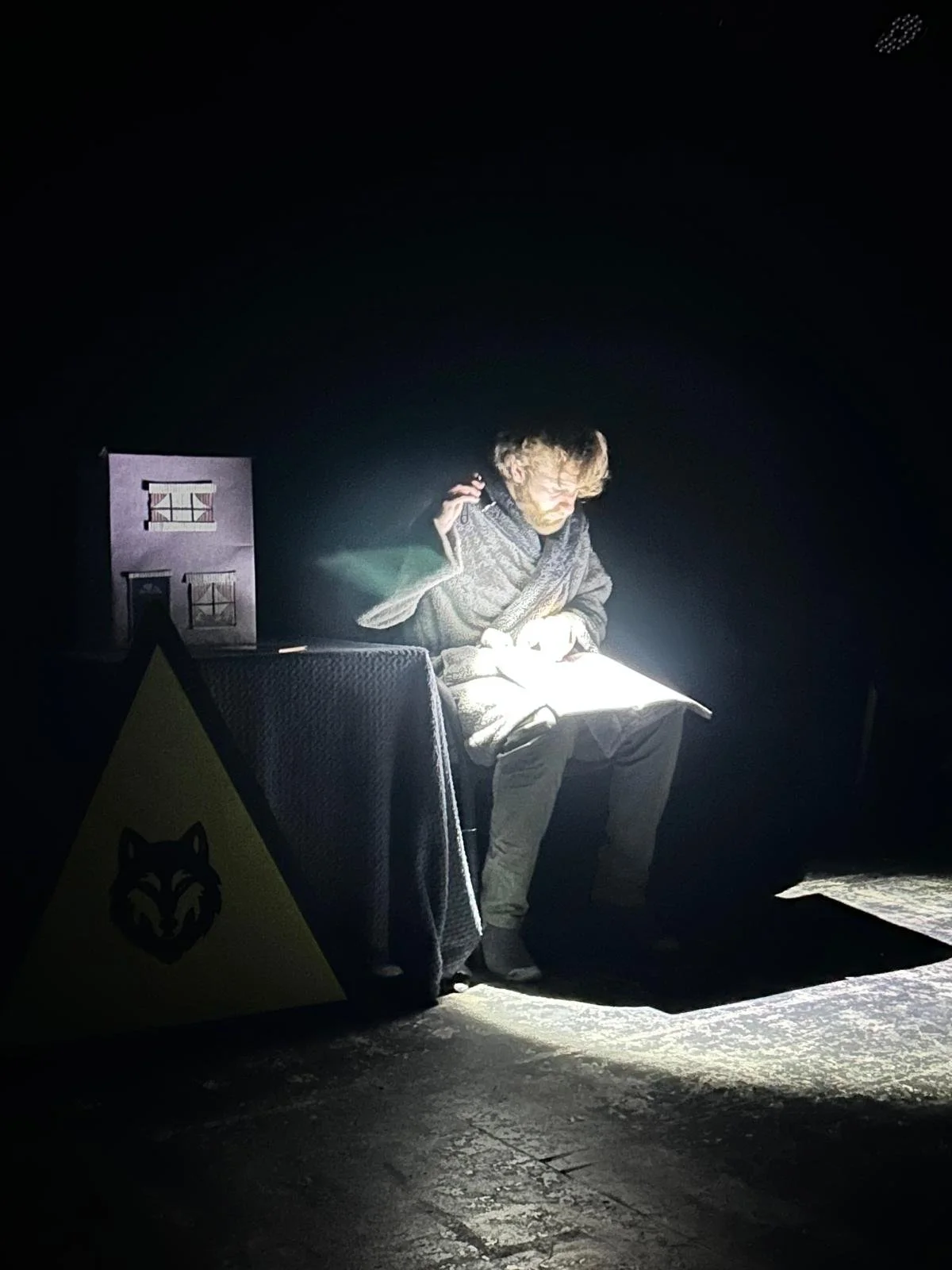Person sitting on stage reading a book, illuminated by a light, with a small house model and a yellow sign with a wolf logo nearby.