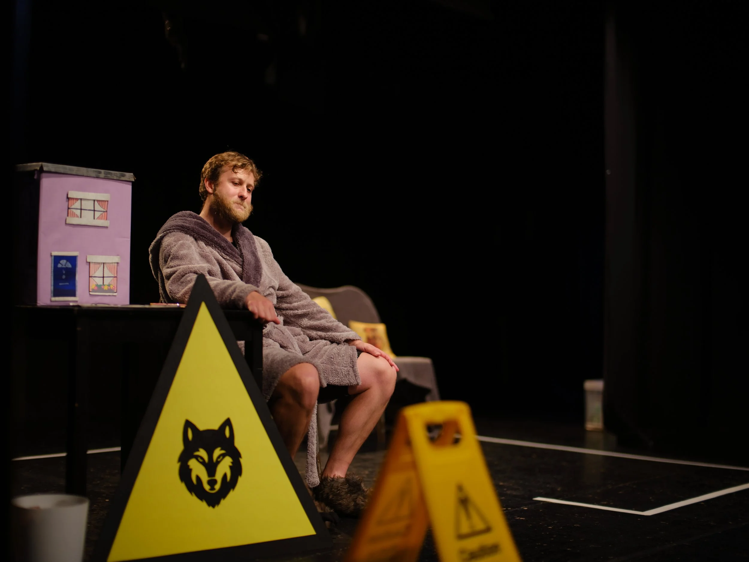 A man with a beard sitting on a chair in a dark room, wearing a bathrobe, with stage props including a pink toy building and yellow caution signs in front of him.