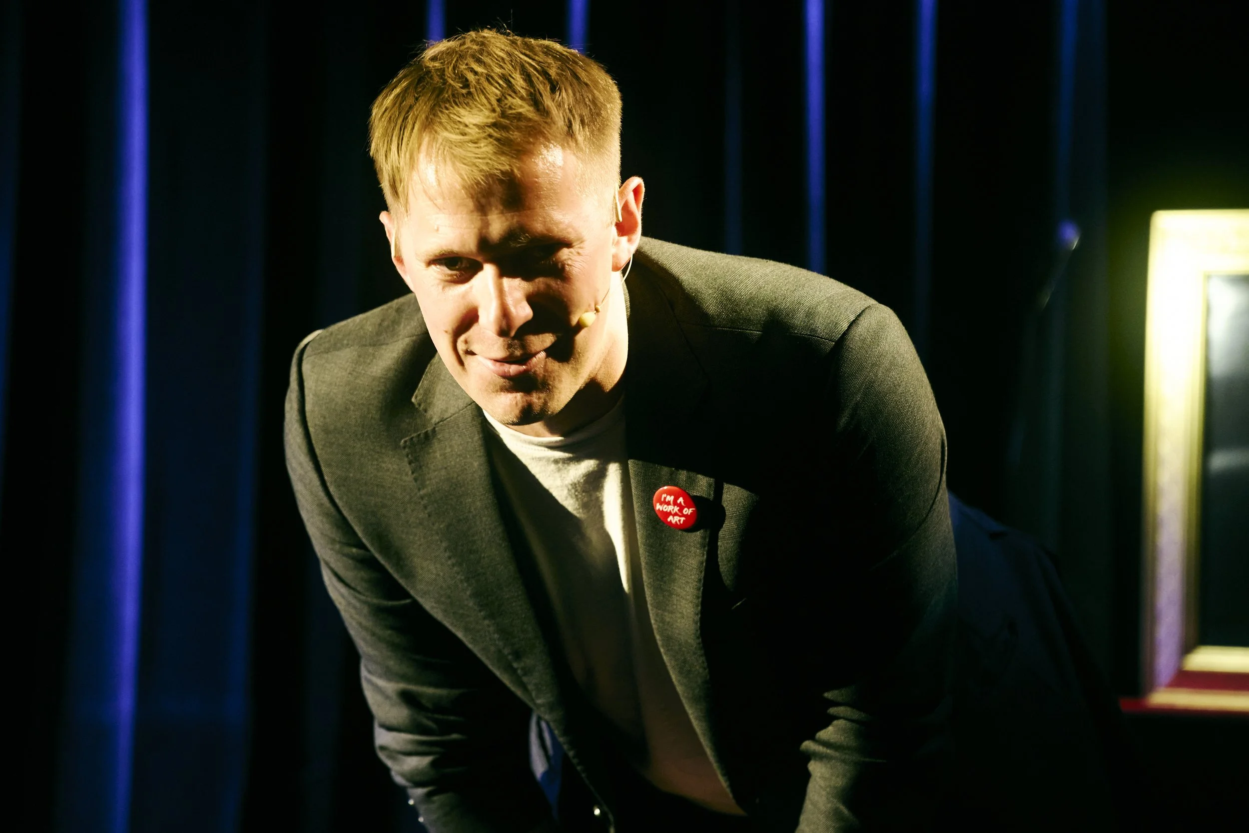 A man with short blonde hair wearing a dark blazer and a button that reads 'I'M A WORK OF ART' is leaning forward on a stage with dark curtains in the background.