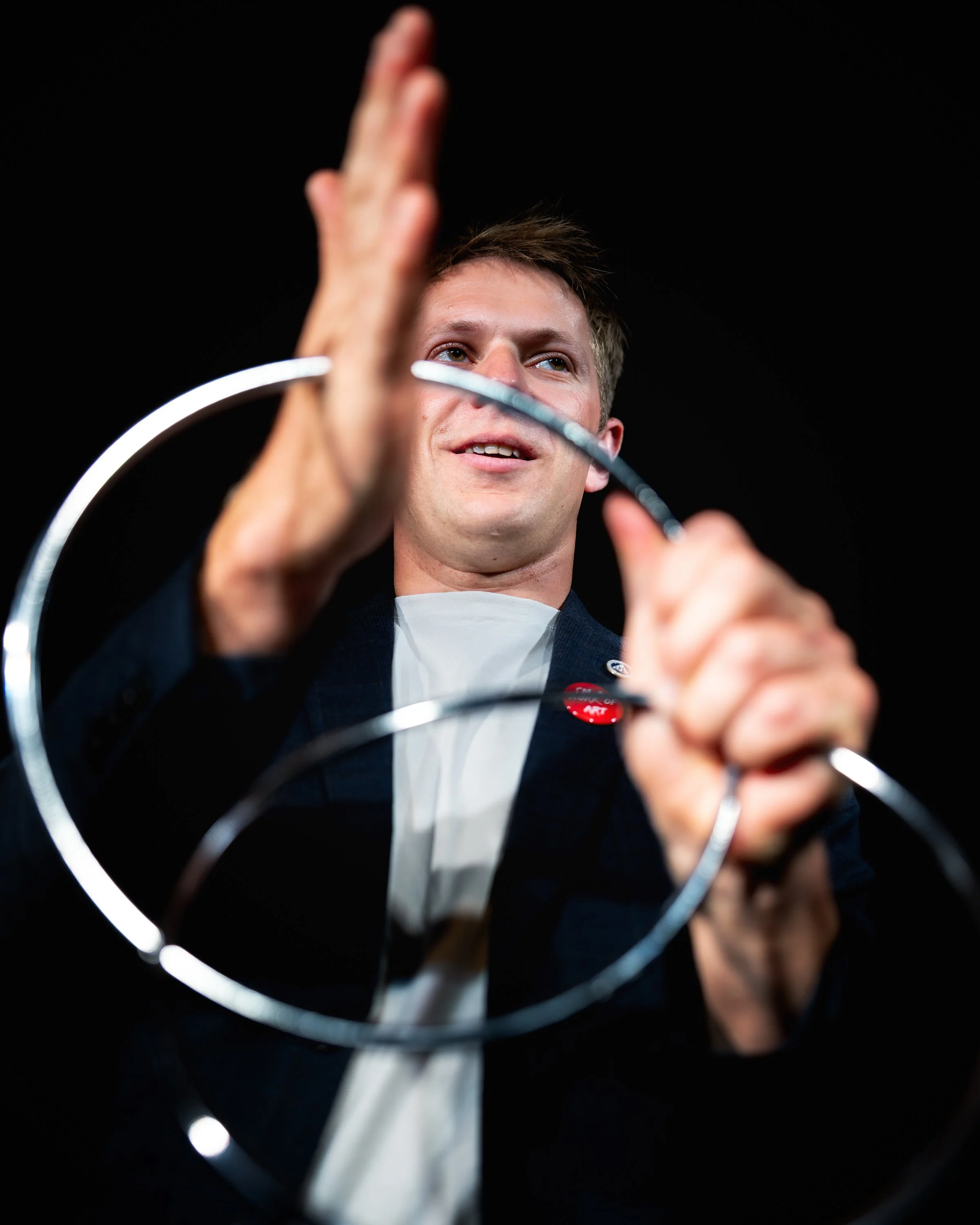 A young man with short brown hair, dressed in a suit and white shirt, is smiling and holding a piece of white string or cable. The background is black, and the photograph is taken from a low angle, focusing on his face and hand.