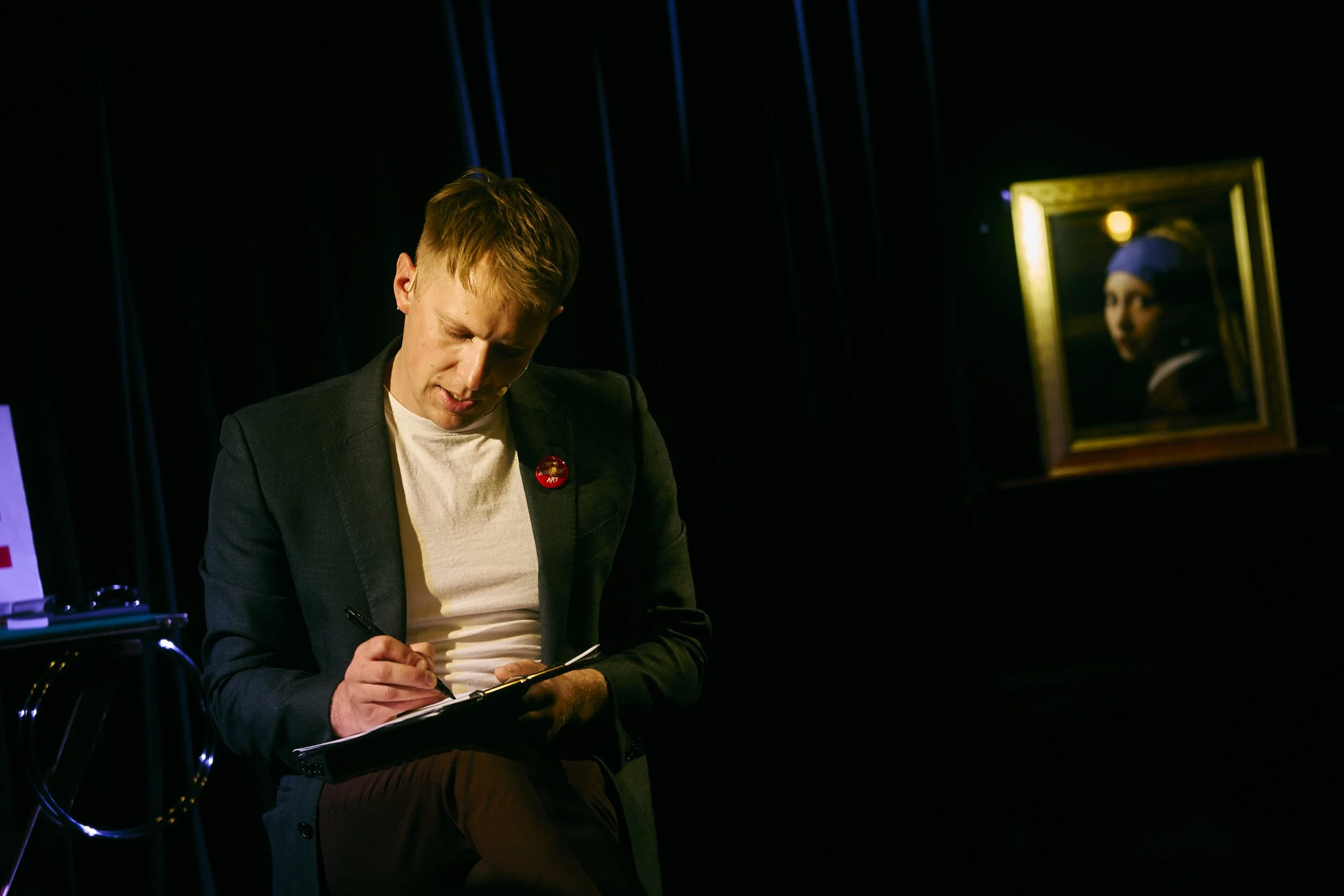 A man sitting on a stool, writing on a clipboard in a dark room with a painting of a woman in the background.