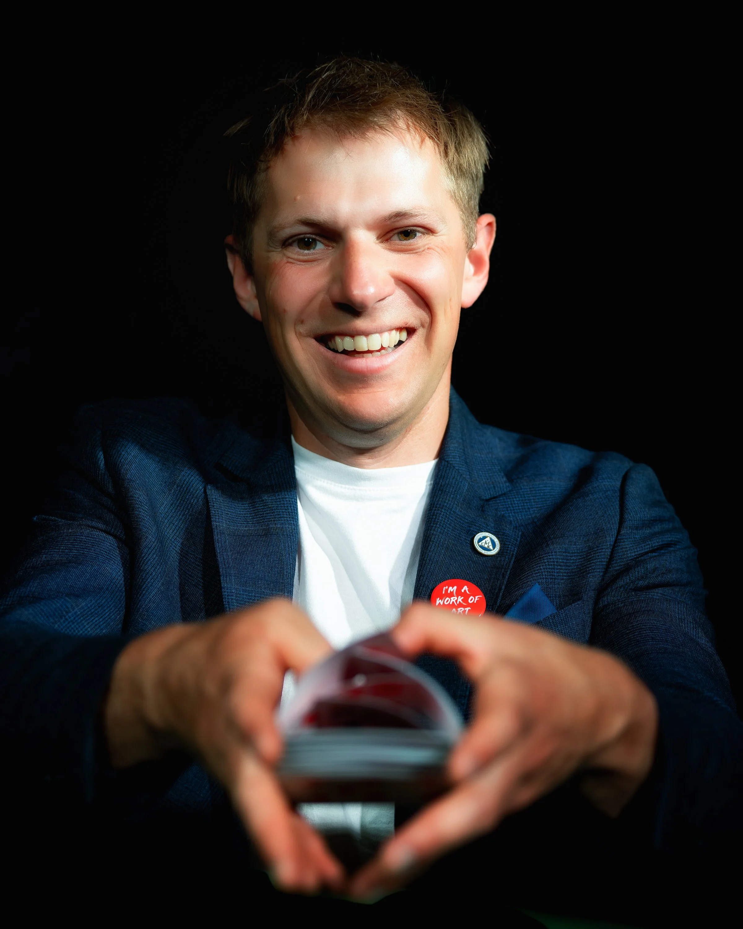 A man in a blue blazer smiling broadly, holding a card with his hands forming a heart shape. He is wearing a red button that says 'I'm a work of art' and a small pin on his blazer.
