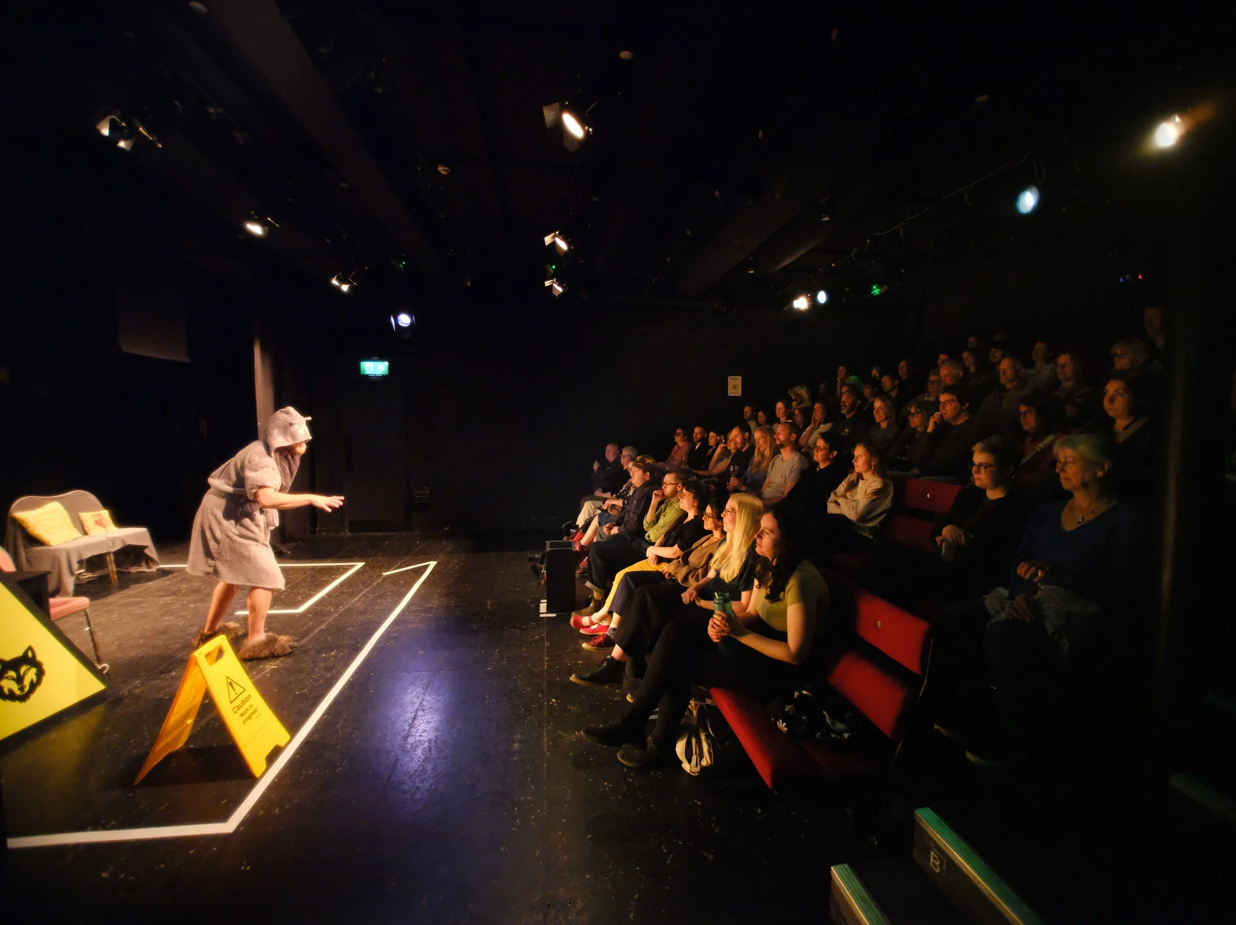 Audience watching a theater performance with a person on stage dressed in costume and wearing a furry cap. The audience is seated in rows with some wearing glasses, and there are warning signs on the floor.