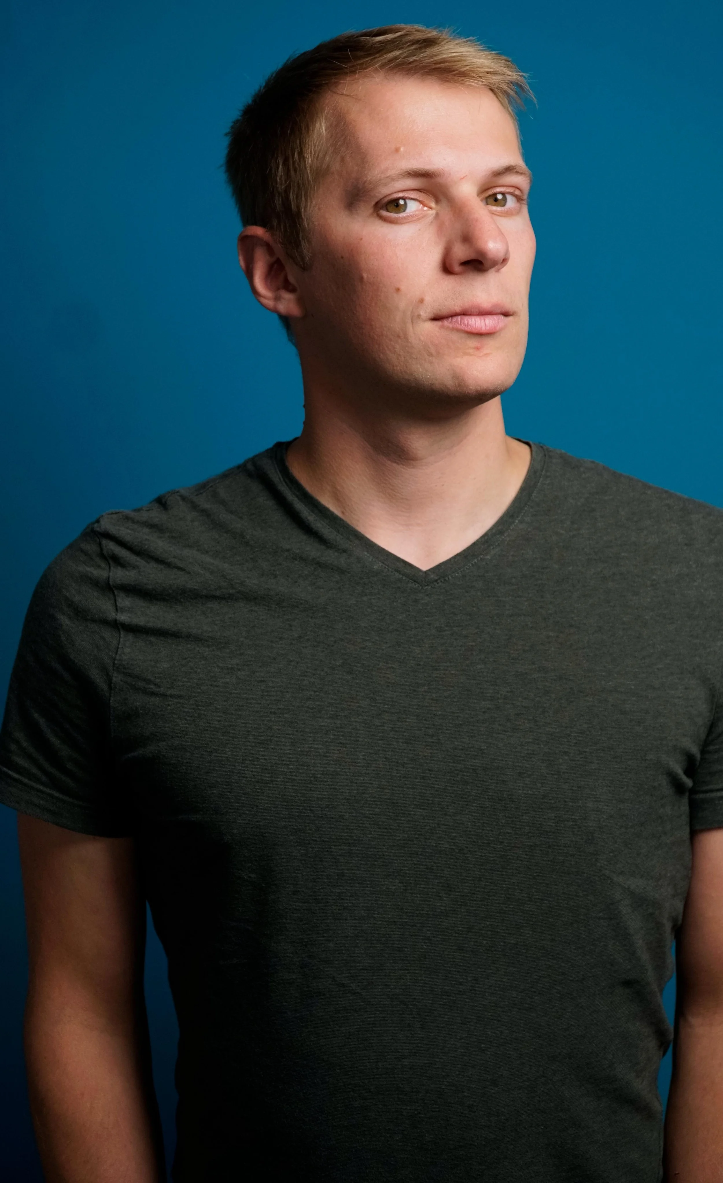 A young man with short, light brown hair and hazel eyes wearing a dark gray V-neck T-shirt, standing against a solid blue background.