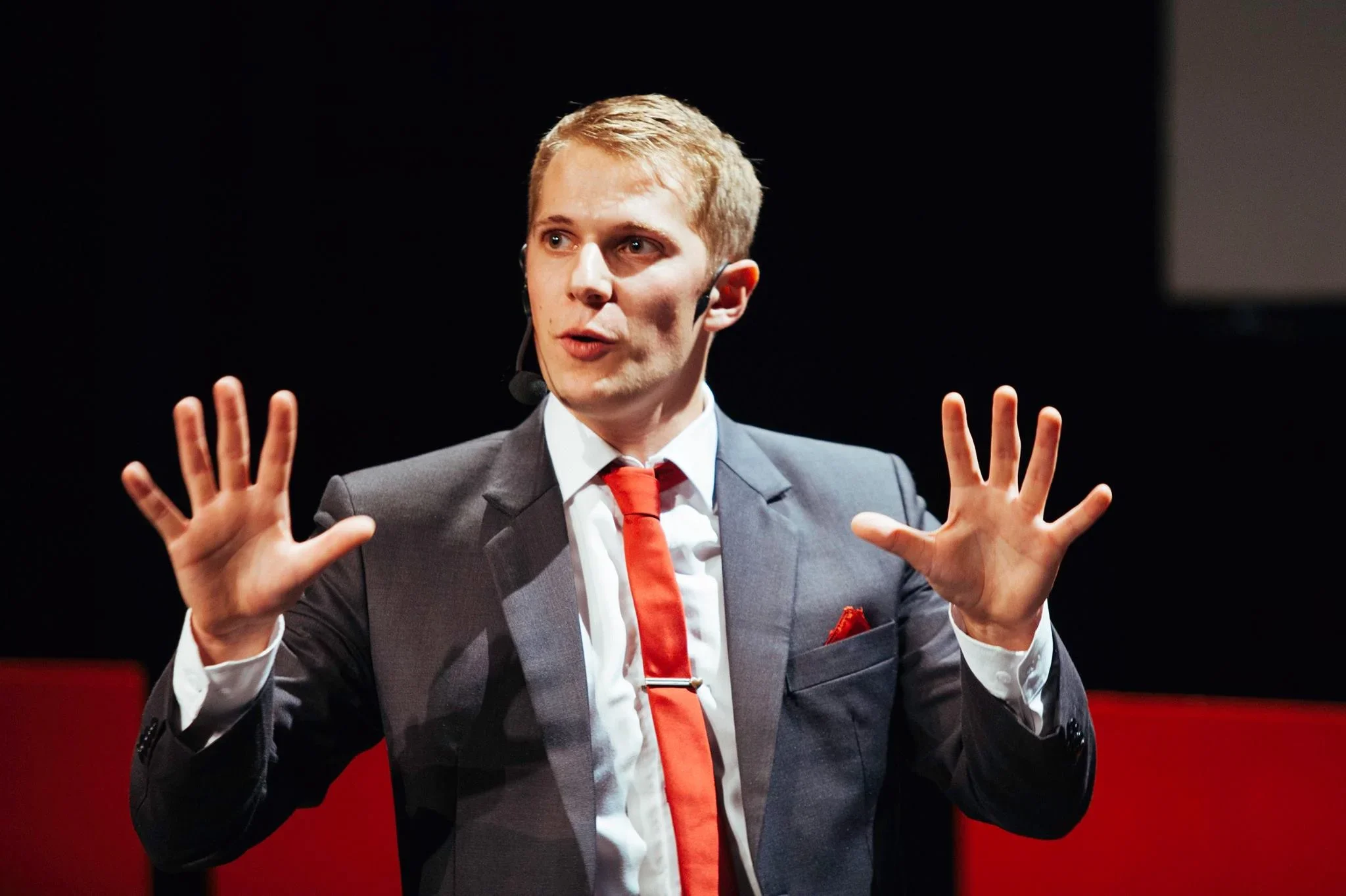 A man in a suit with a red tie and pocket square is speaking or presenting on stage, wearing a headset microphone with black ear pieces, against a dark background with a red stripe.