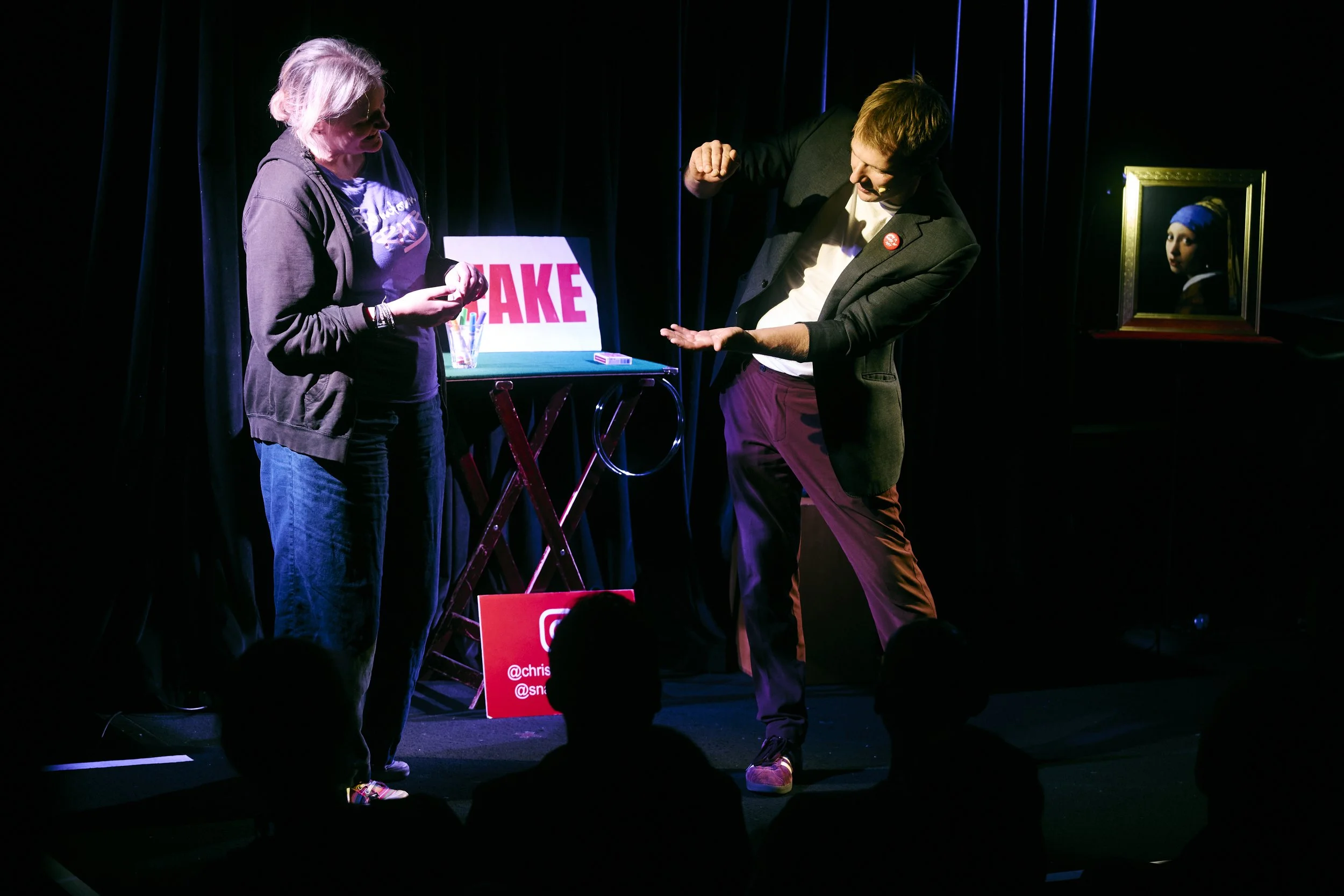 A woman and a man performing on a black stage with a dark background and curtains. The woman is holding a small object, and the man is showing his open palm. There is a table with a sign reading "FAKE," a glass of water, and some items on it. A frame