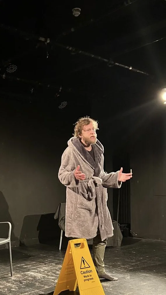 A man with curly hair and a beard performing on a black stage, wearing a gray bathrobe tied at the waist. A yellow caution sign indicating work in progress is on the floor in front of him. The stage has black walls and a few chairs, with stage lighti