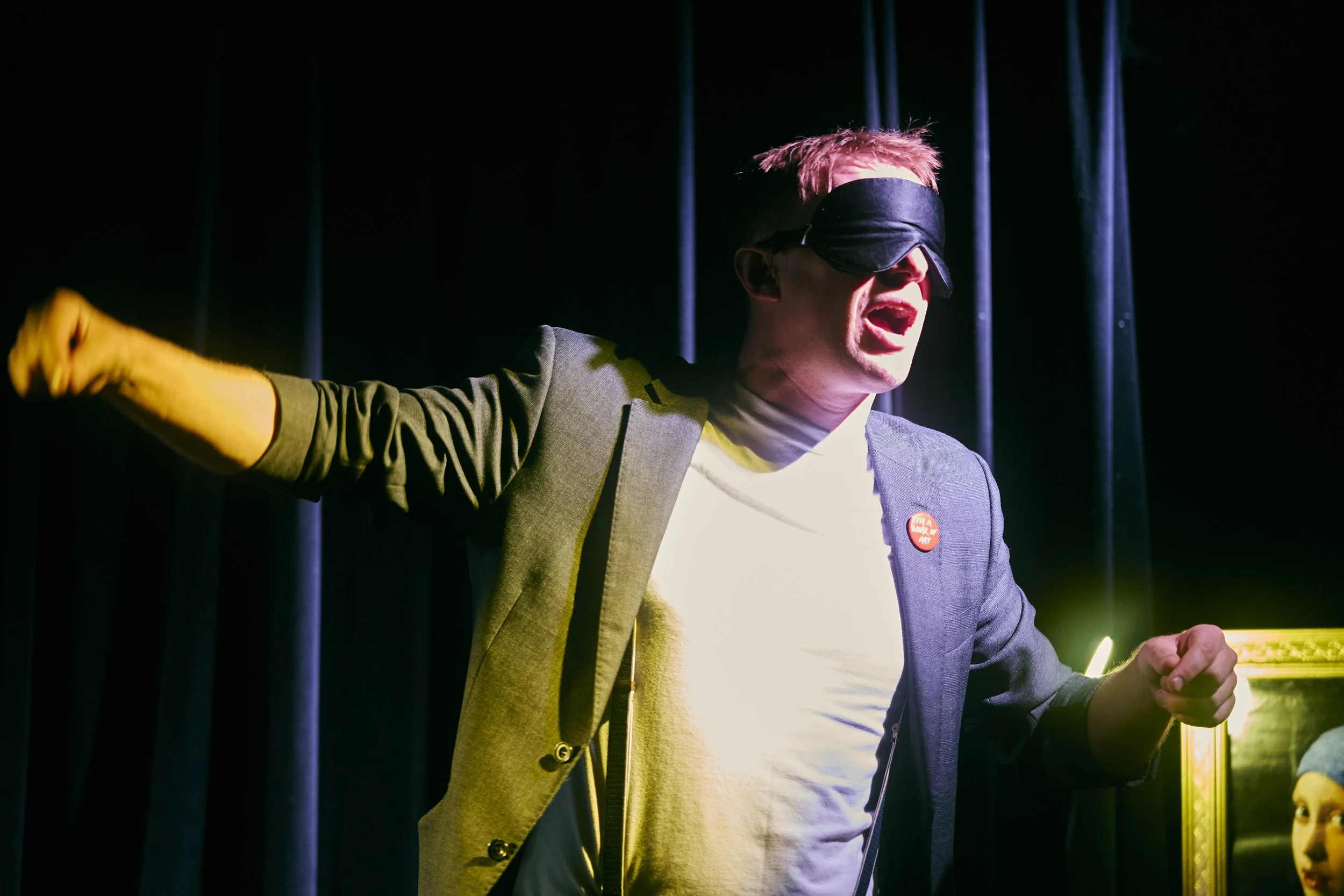 Person wearing a suit with a red pin, blindfolded, and with mouth open, appears to be shouting or singing, with a curtain and light background.