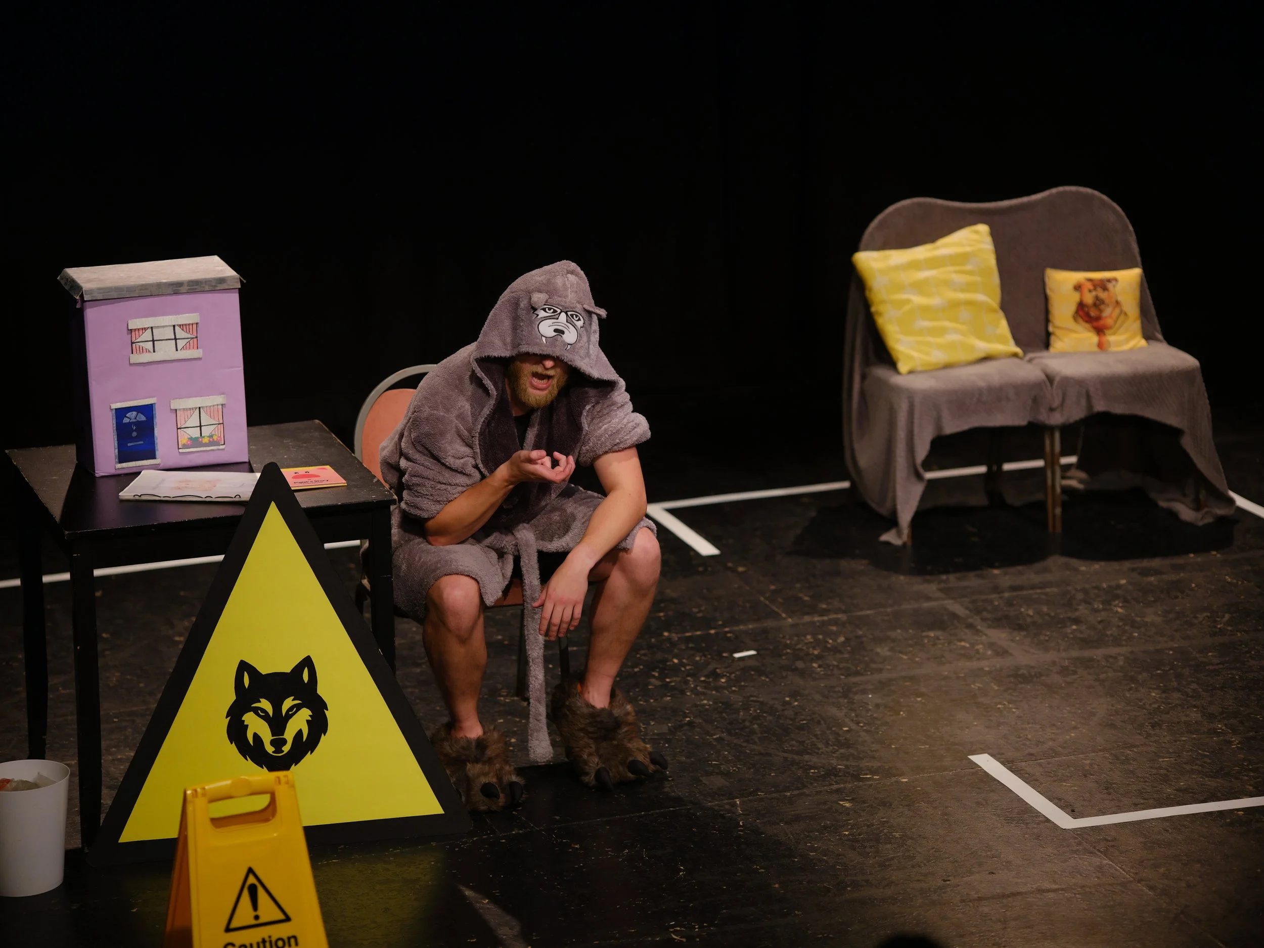 Person dressed in a bear costume with bear paws, seated on a chair, in a theatrical setting with signs, a dollhouse, and yellow caution signs nearby.