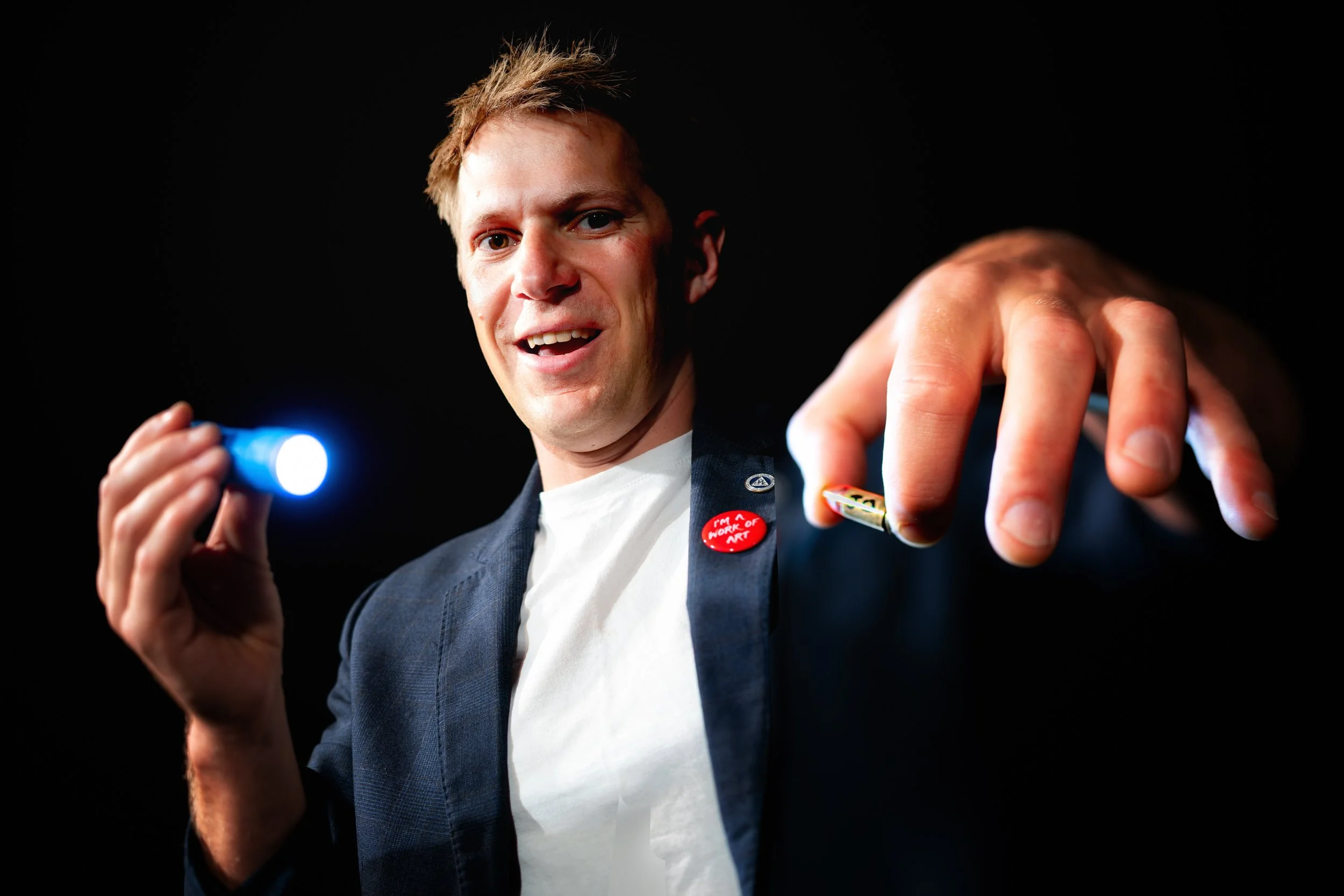 A man with light hair pointing a flashlight at the camera, wearing a blazer and a white shirt, against a black background.