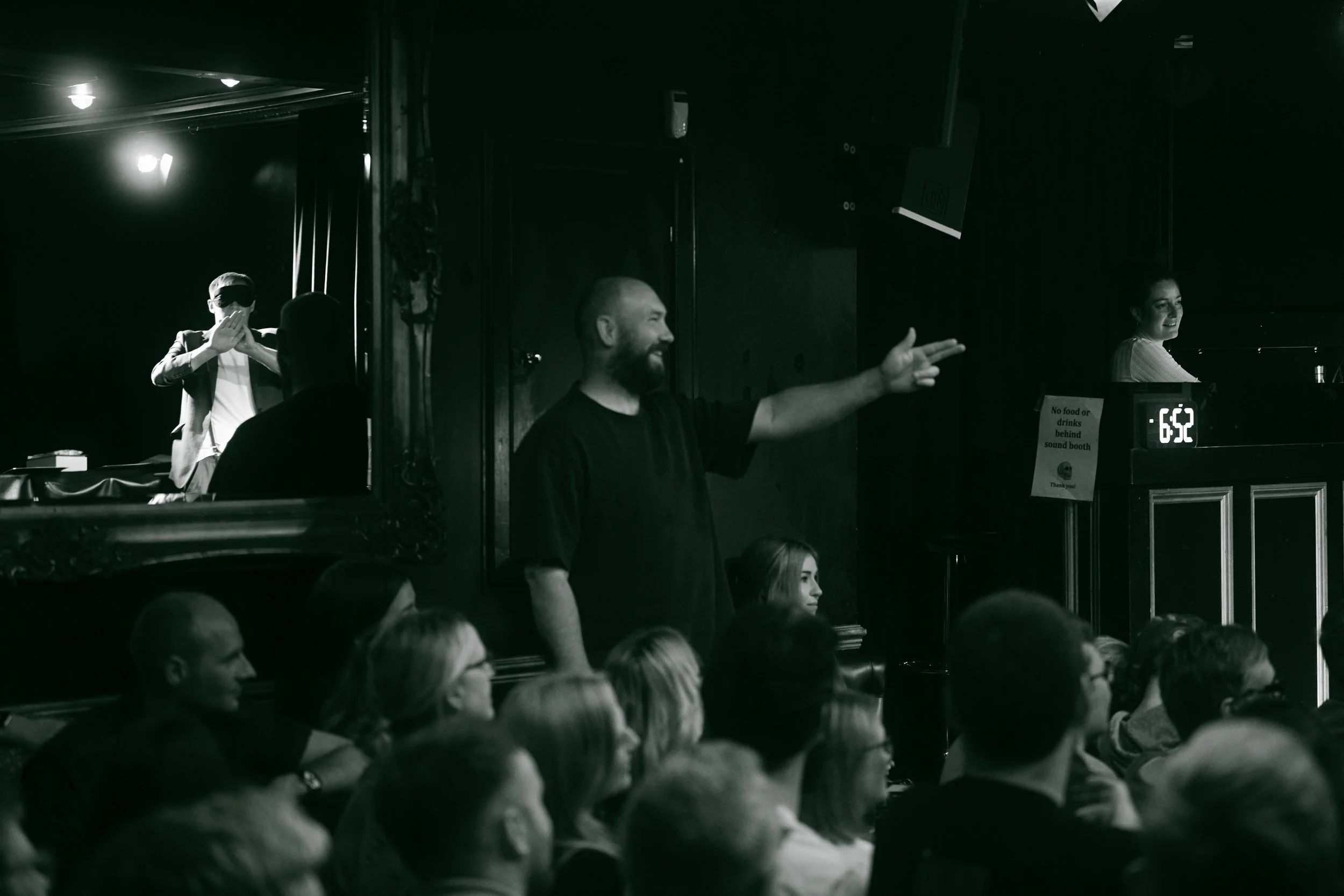 A man with a beard and a black shirt is standing among an audience, smiling and gesturing with his right hand. Behind him, there is a woman at a piano and a sign that says 'No food or drinks behind sound booth.' The scene appears to be at an event or