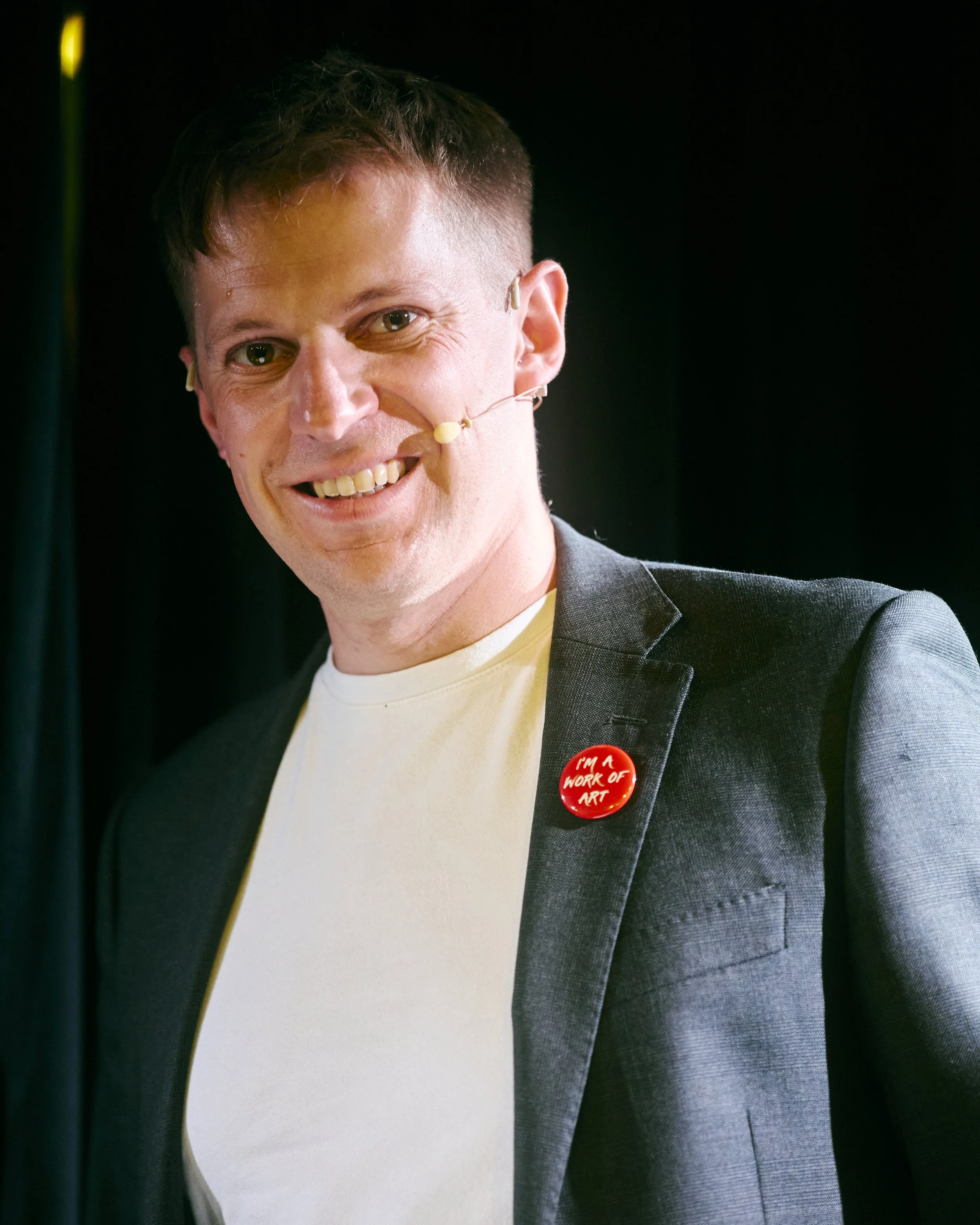 Close-up of a smiling man wearing a grey blazer, a cream-colored shirt, and a red button that says 'I'M A WORK OF ART'. He has short brown hair, light skin, and a flesh-colored microphone headset.