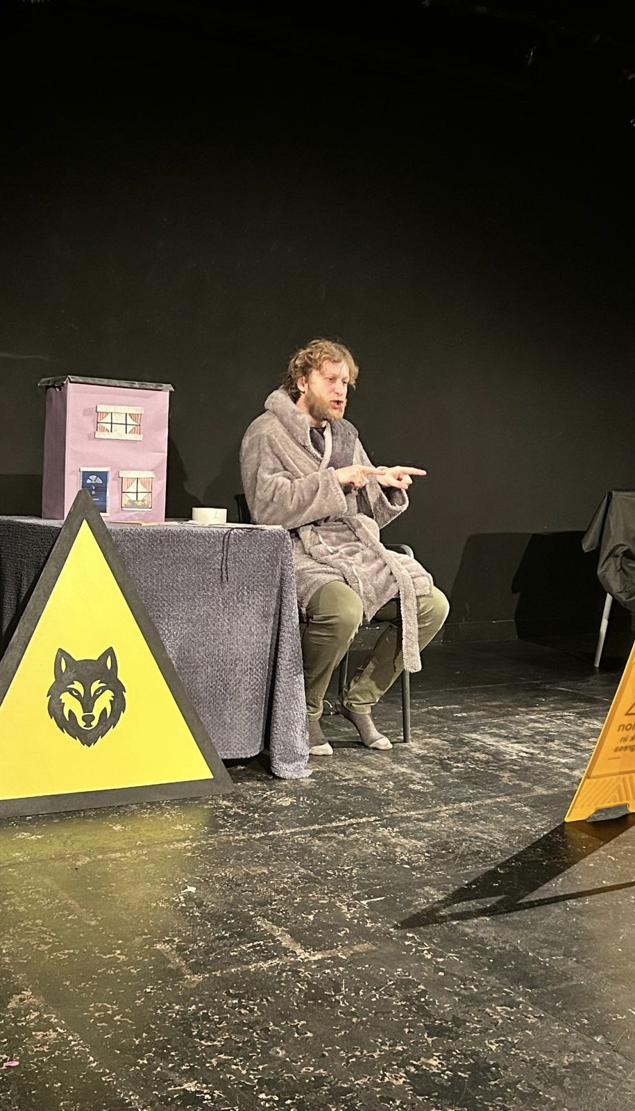 A man in a gray robe appears to be performing or acting on stage, seated in front of a black background. There are signs with a wolf illustration and a caution sign near him, and a table with a pink box and a white cup.