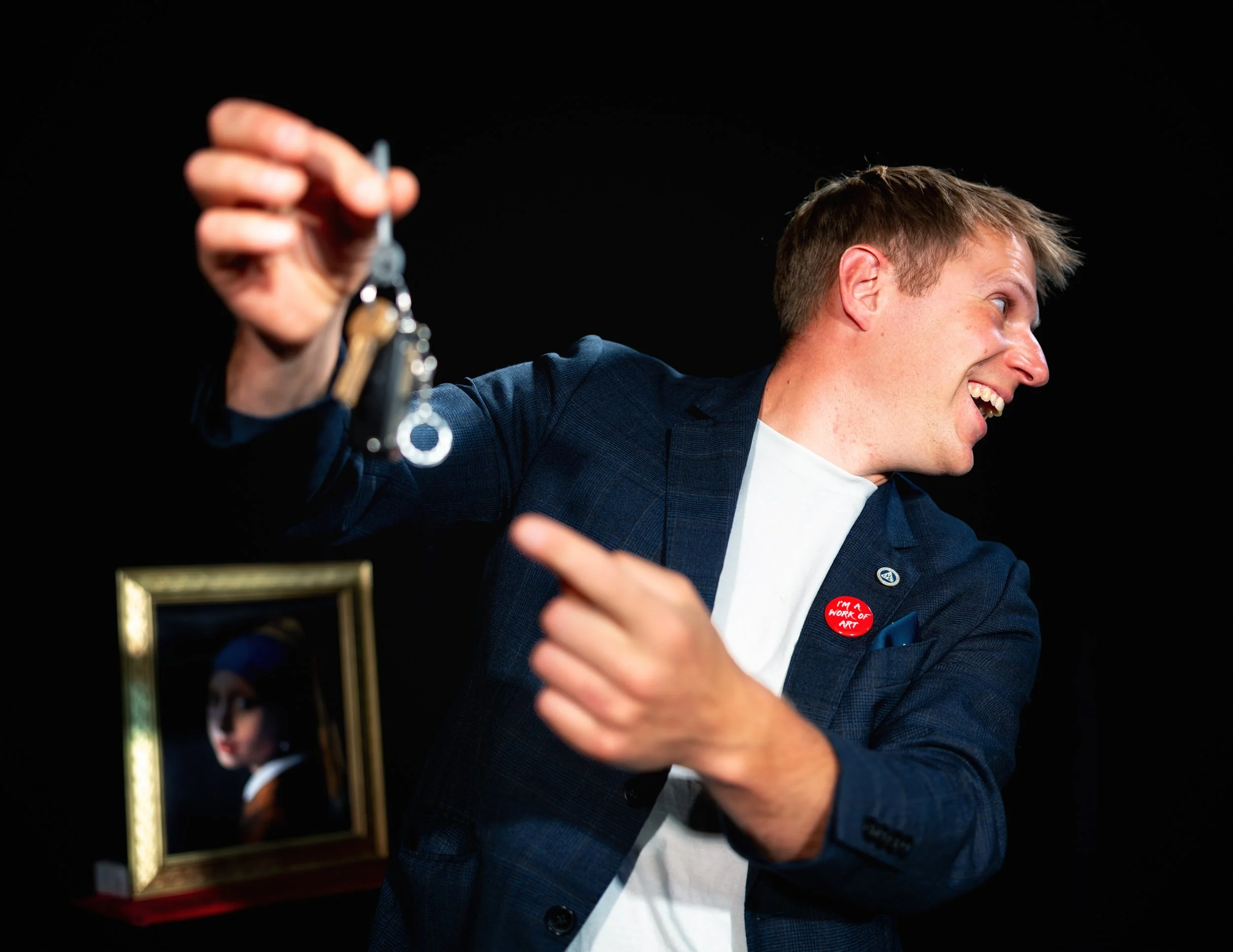 A man in a blue suit holding a set of keys close to his face, smiling, with a woman in the background and a gold-framed mirror on a red table.