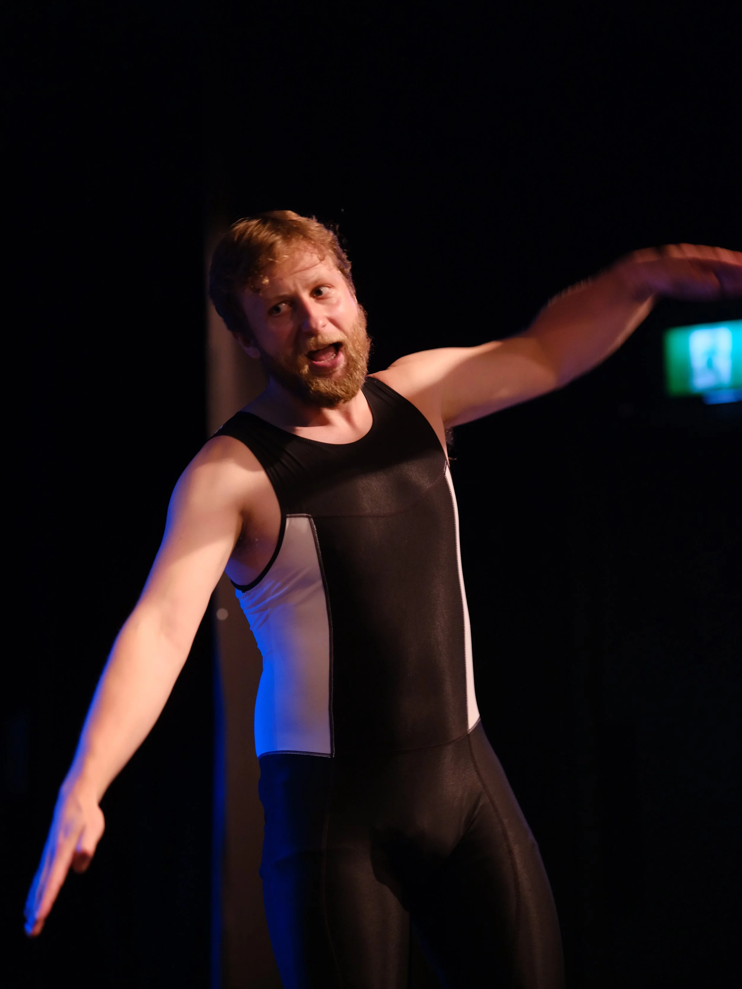 A man with red hair and beard wearing a black and gray athletic tank top and black pants, with his arm extended outward, in a dark setting with subtle blue and purple lighting.