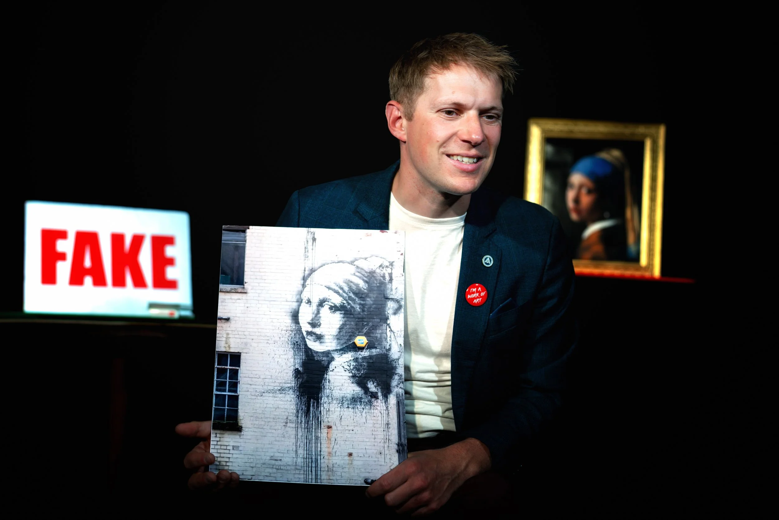 A man with short light brown hair smiling, holding an artwork of a black and white portrait of a woman on a brick wall. In front of him, there is a sign reading "FAKE" in red letters, and he wears a button that says "I'M A WORK OF ART." A framed pain