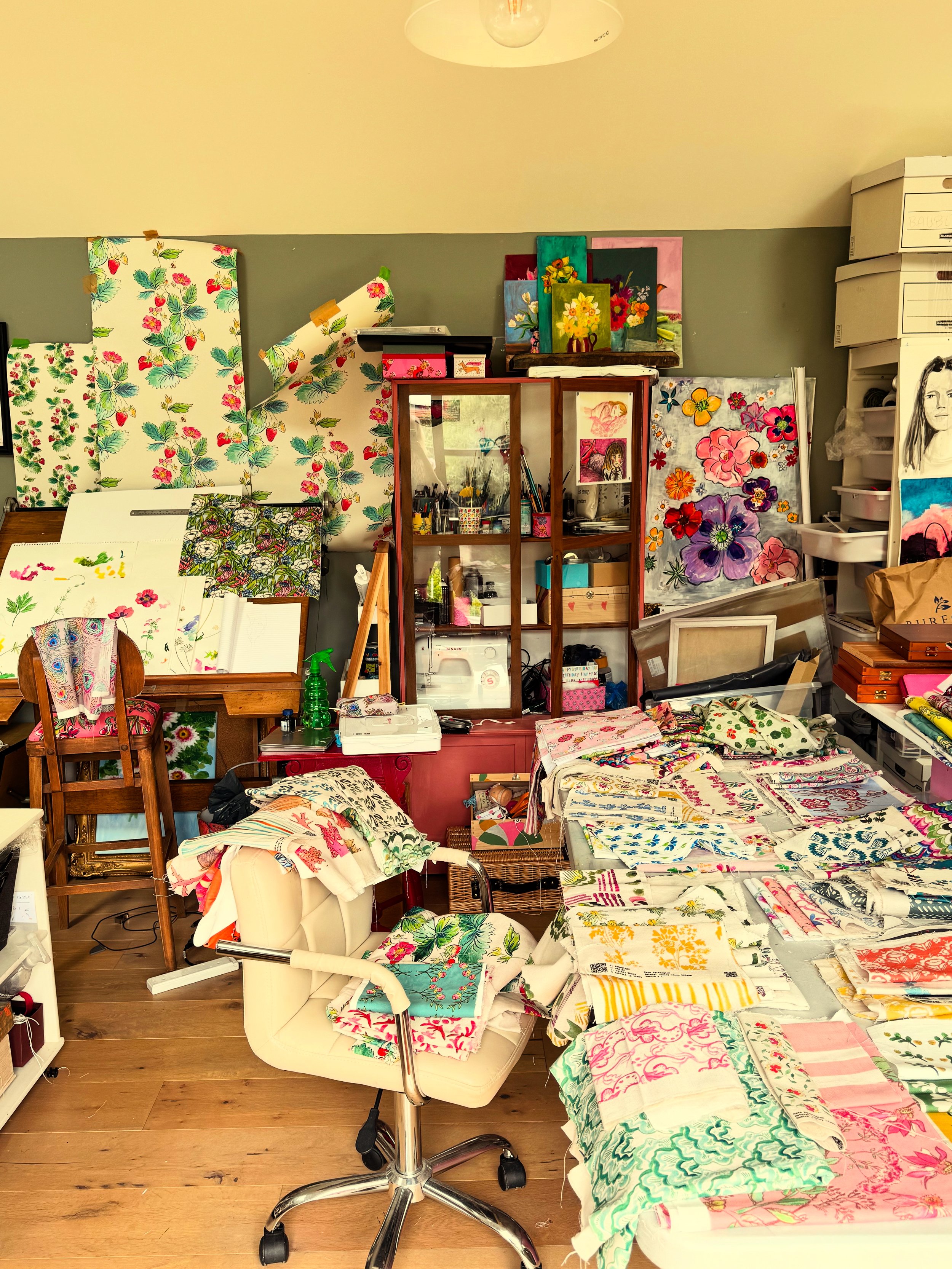 A craft or art studio with various floral fabric prints and paintings displayed on the walls. There are tables covered with fabric samples, a glass cabinet with art supplies, and a white armchair with fabric draped over it. The room has a wooden floo