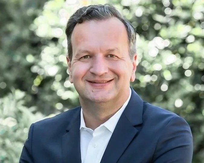 Ein Mann in einem dunkelblauen Anzug und weißem Hemd, lächelnd vor einem Hintergrund aus grünen Bäumen und Licht, im Freien.