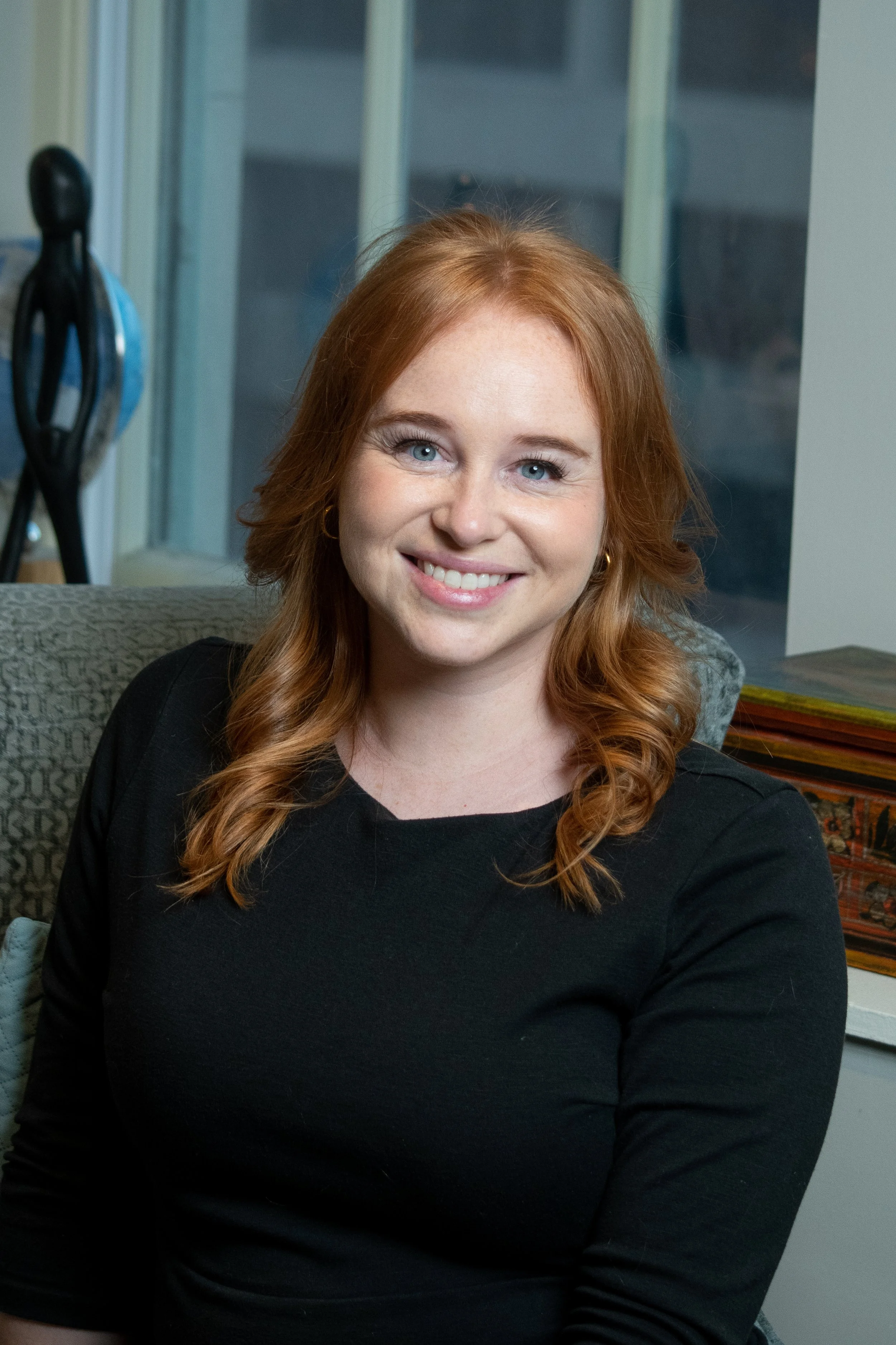 A young woman with red hair and blue eyes, smiling and wearing a black top, sitting in an indoor setting.