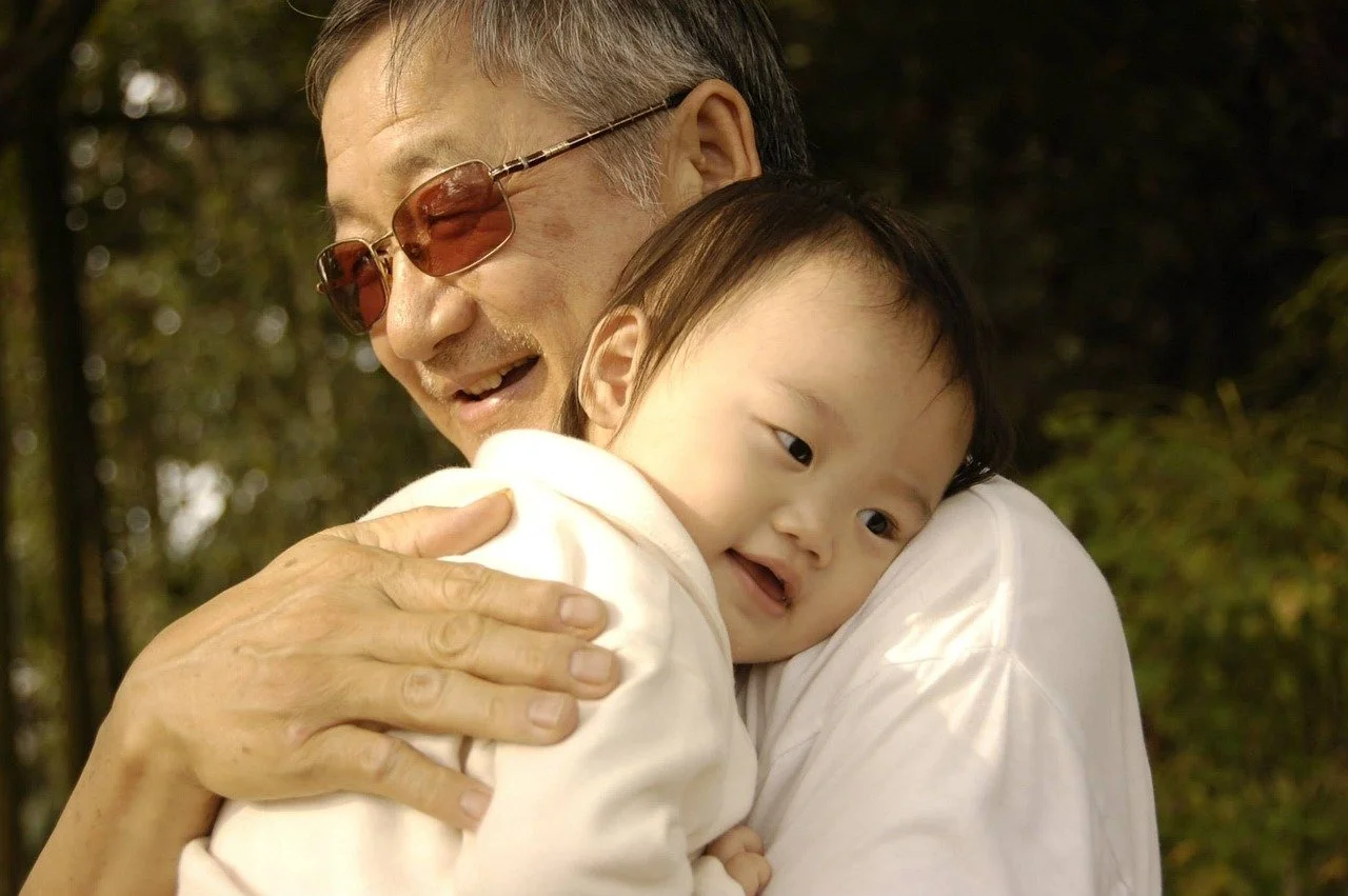An elderly man with sunglasses hugging a young child, both smiling, outdoors with trees in the background.