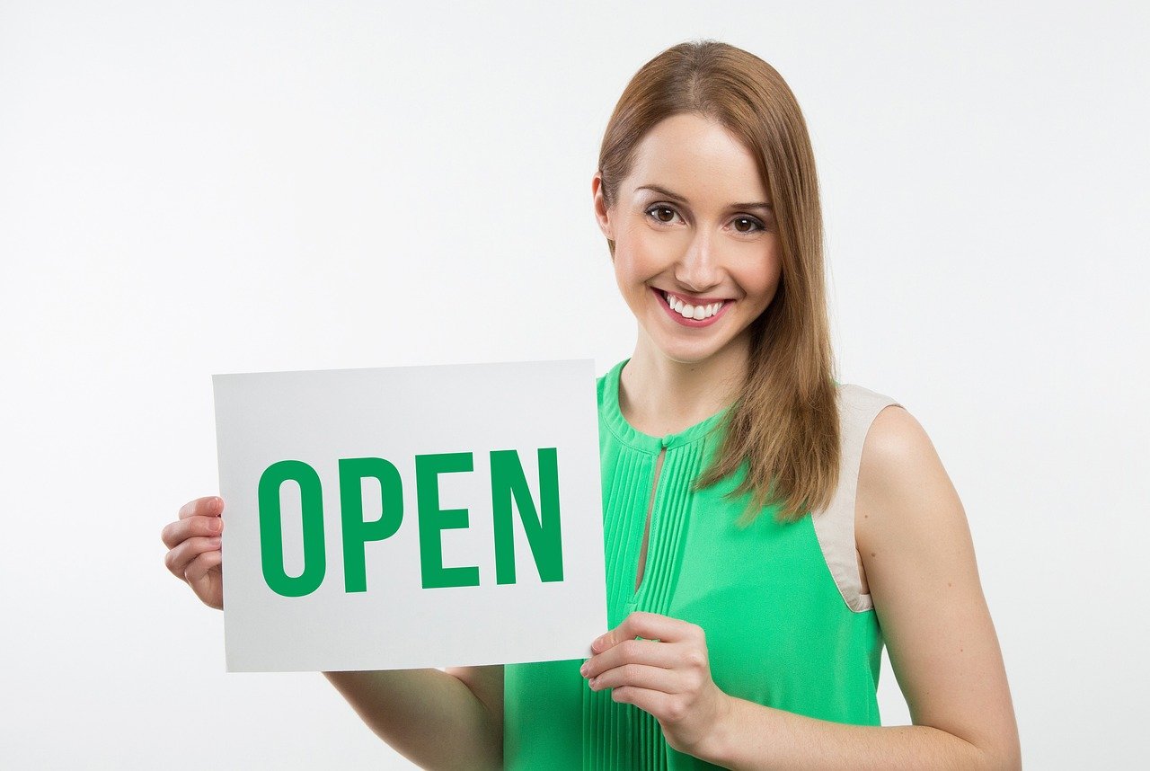 A woman smiling and holding a sign that says 'OPEN' in green letters.
