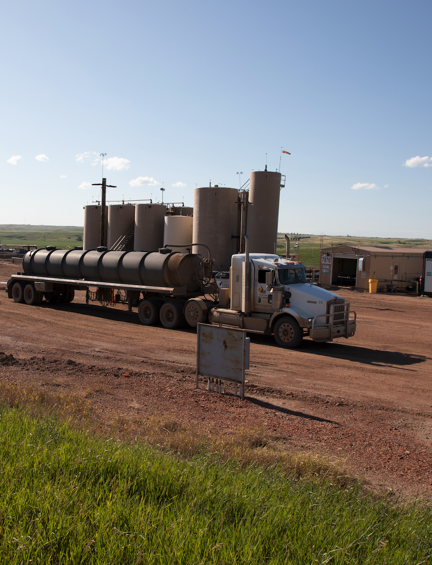 A large white semi-truck with a cylindrical tank trailer parked on a dirt road in a rural area with flat fields and blue sky in the background.