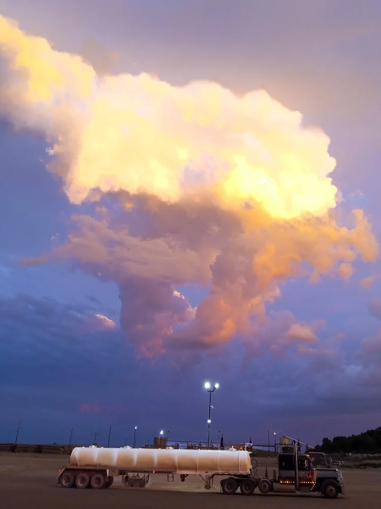 A large truck with a tank trailer parked outdoors under a colorful sky with large, illuminated clouds.