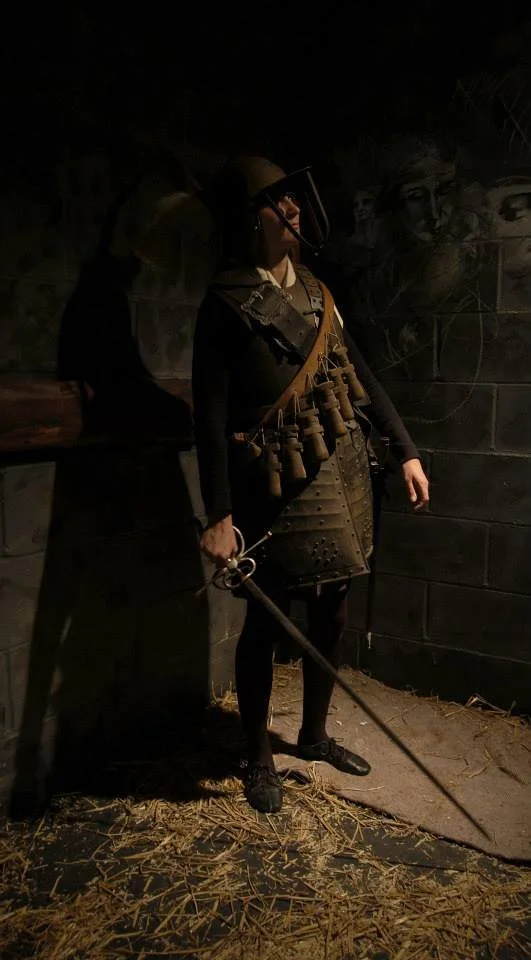 A person dressed in medieval or fantasy armor, holding a sword, standing in a dimly lit room with mural art on the walls.