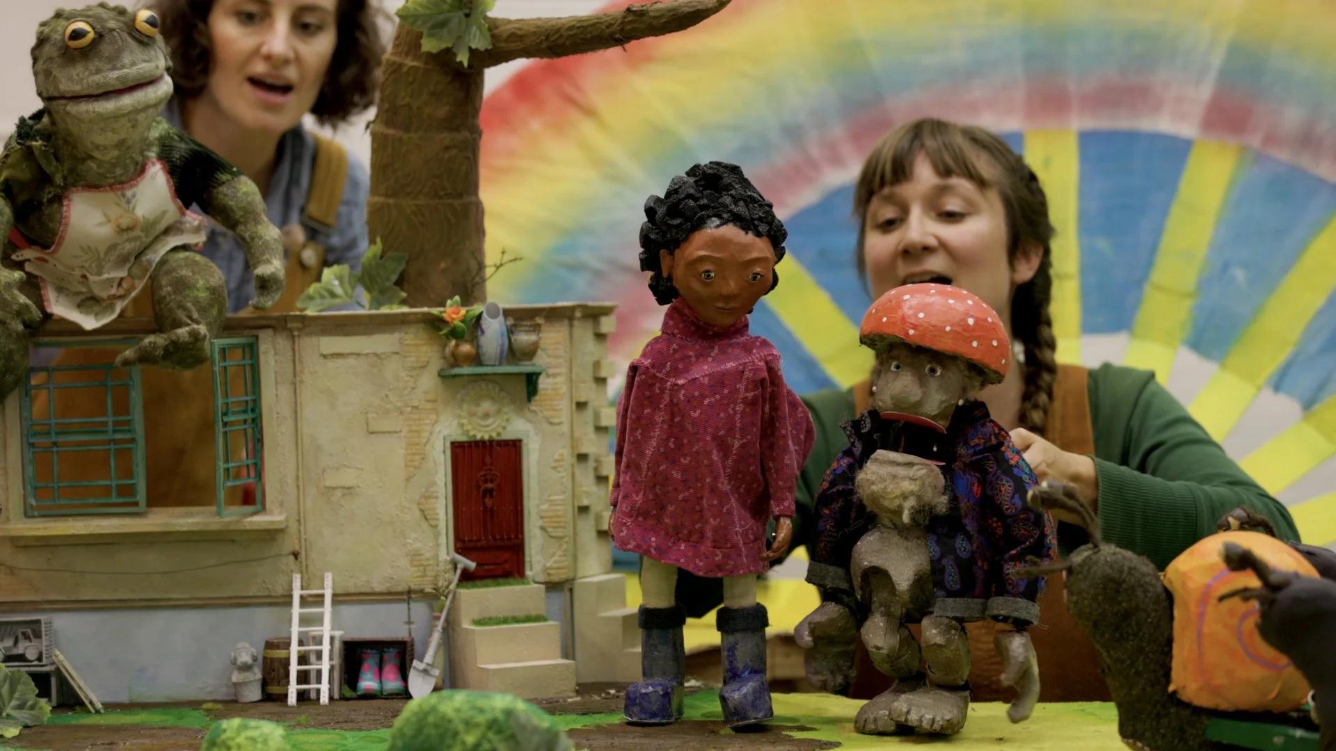 Two children and two puppets in front of a colorful, rainbow-colored backdrop. One child is holding a puppet of a person with curly black hair and a red shirt, while the other is holding a puppet of a mushroom with a face. The children are engaging with the puppets, and a woman is observing.