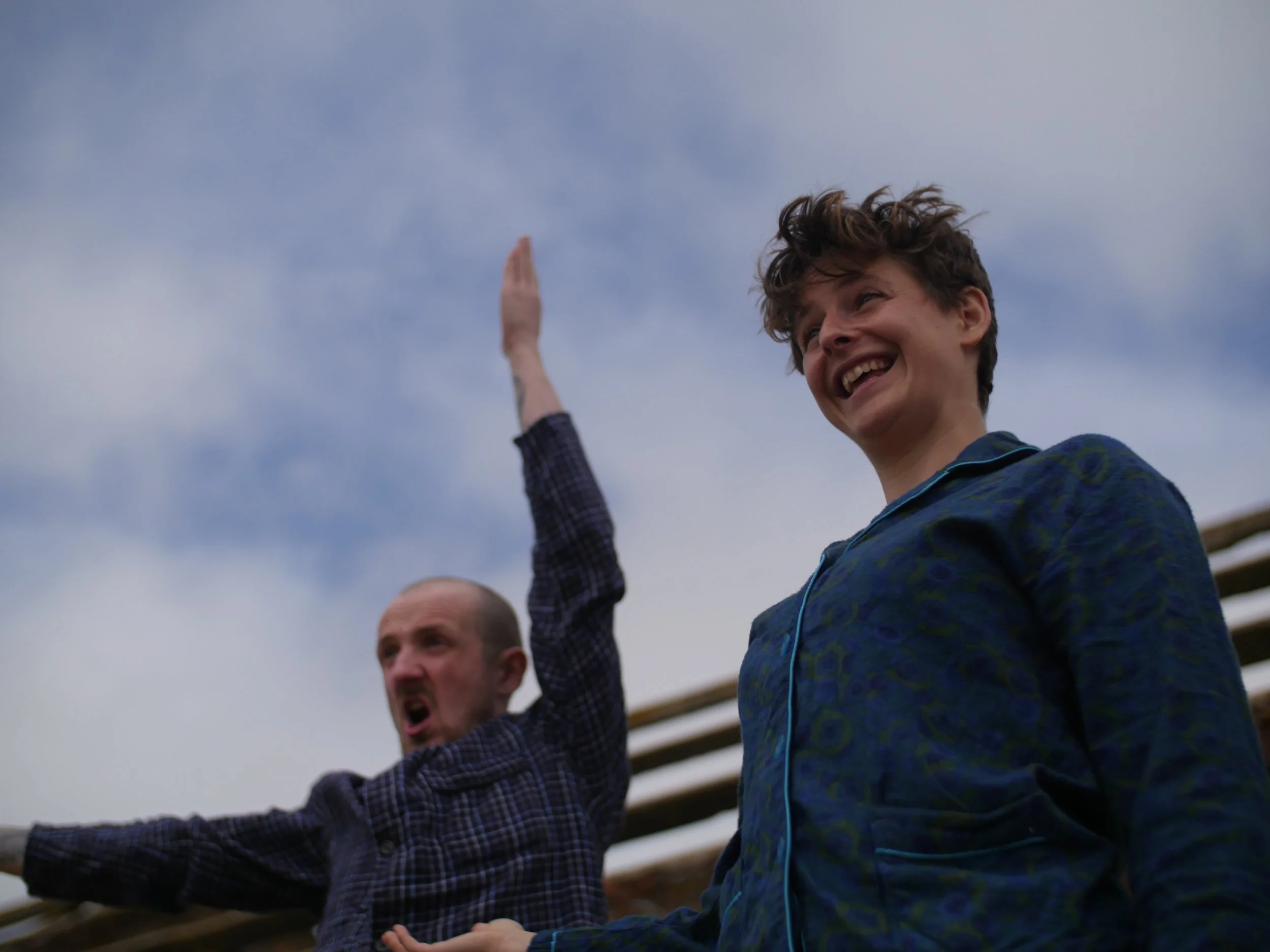 Two young men appear to be cheering or celebrating outdoors, with one smiling and raising his arm, both dressed in casual clothing, against a cloudy sky background.