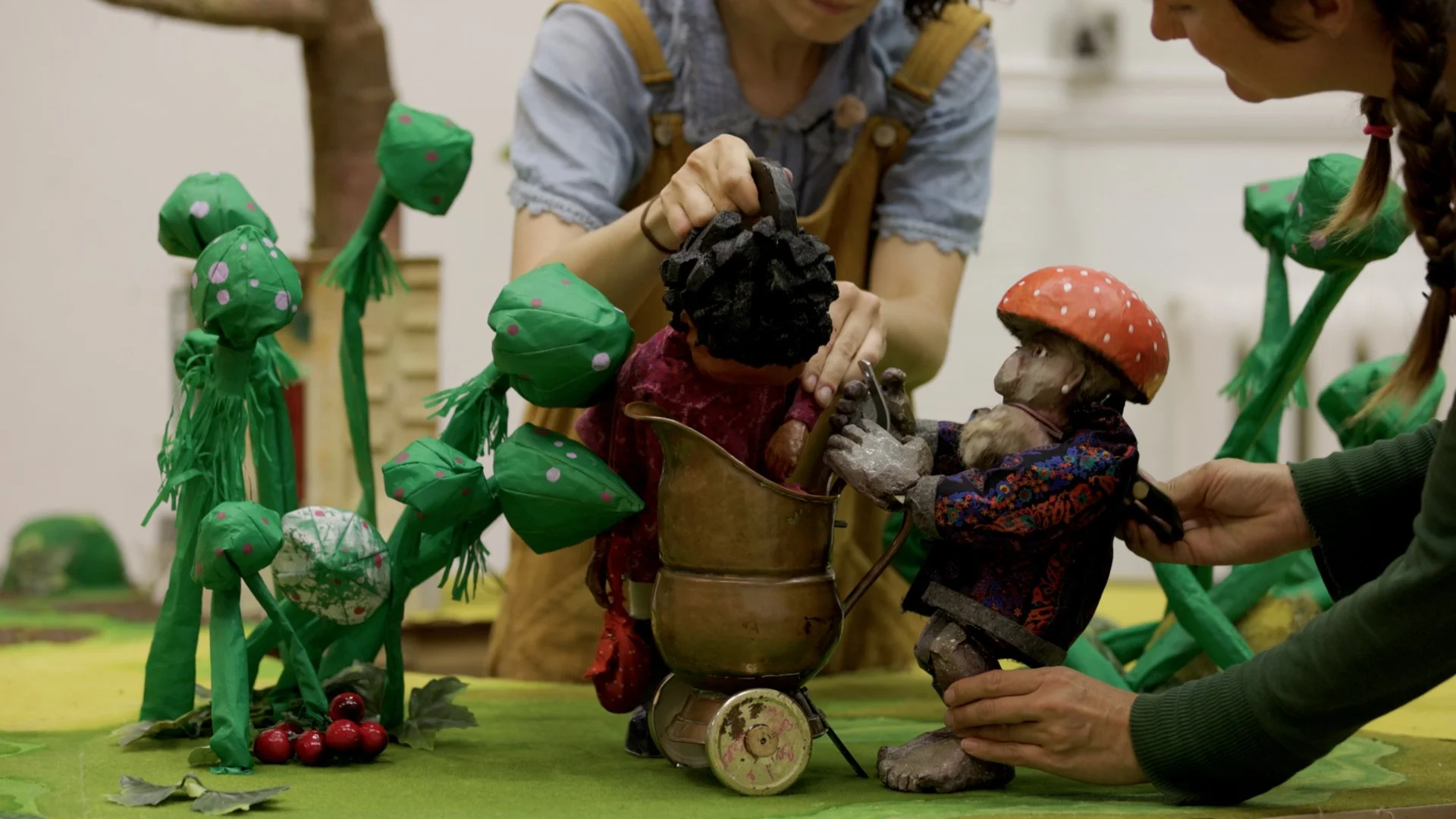 Two people work a puppet girl and a mushroom guy on a green surface.