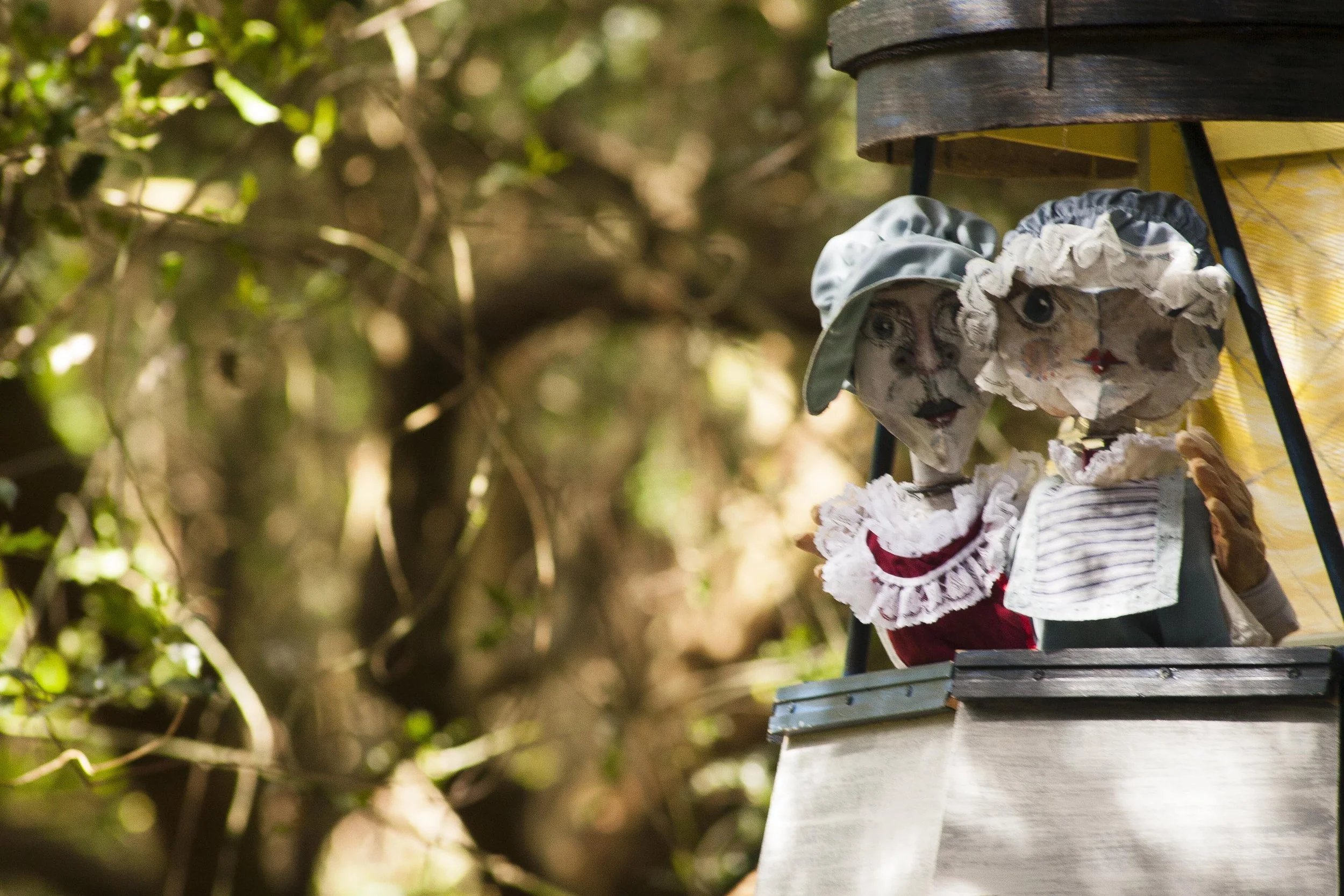 Close-up of a decorative birdhouse with a doll's face and bonnet, mounted on a wooden post with greenery in the background.