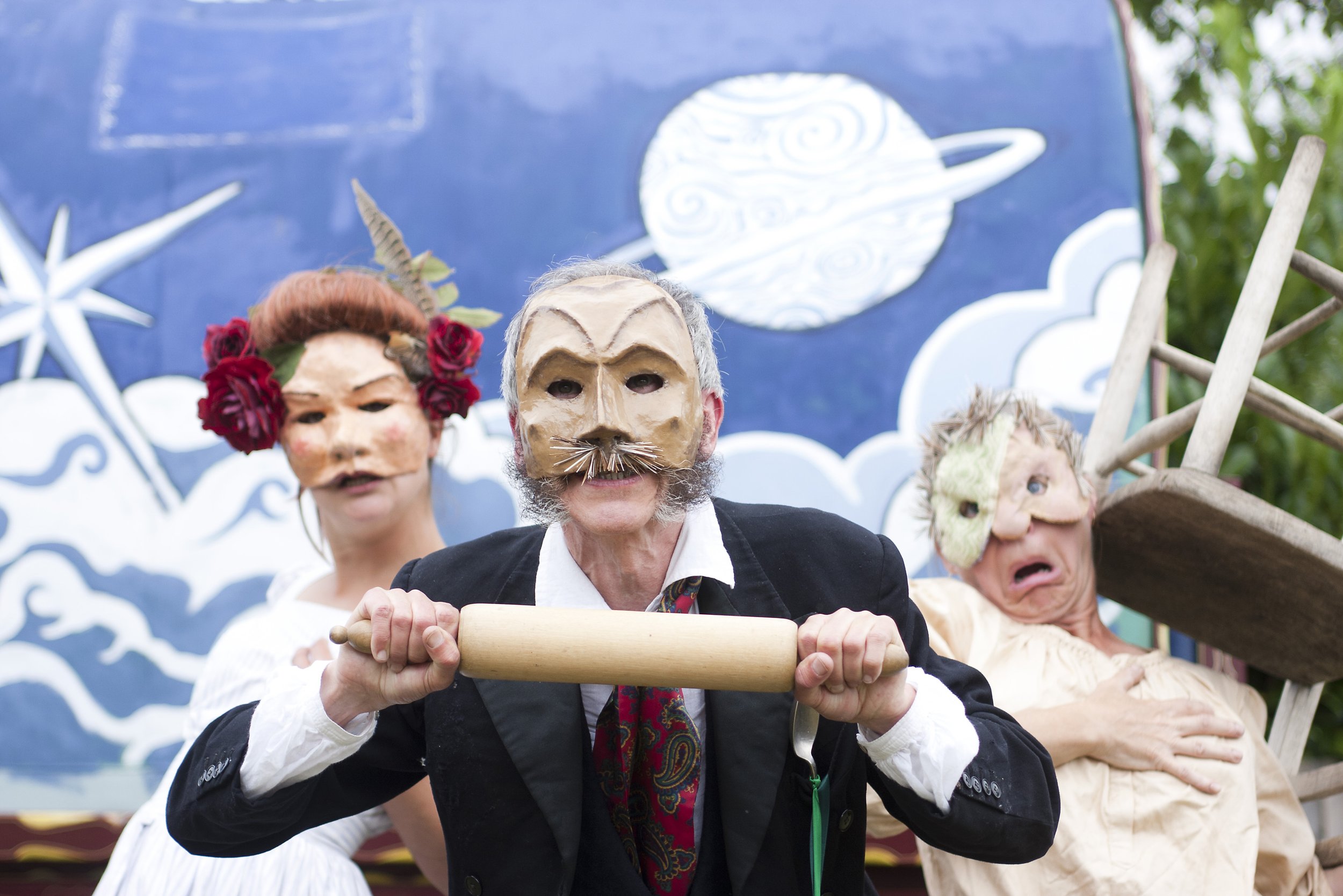 A group of people in masks and costumes participating in a parade, with a blue sky and decorative backdrop featuring a white moon and clouds.
