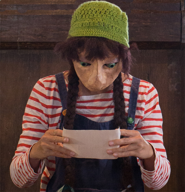 A person dressed as a character from the animated series 'Lupin the Third' with a large nose mask, green knitted hat, red and white striped shirt, and dark overalls, holding a sheet of paper and looking at it.